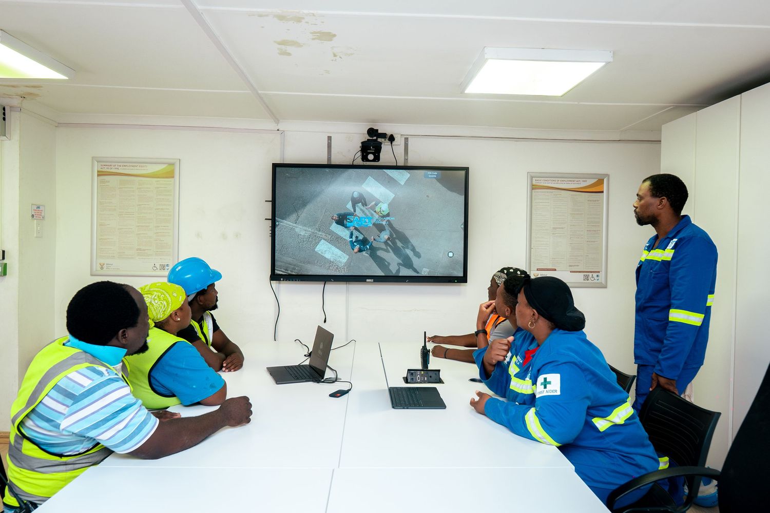 Workers in safety vests watch a training video on a screen during a conference room meeting.