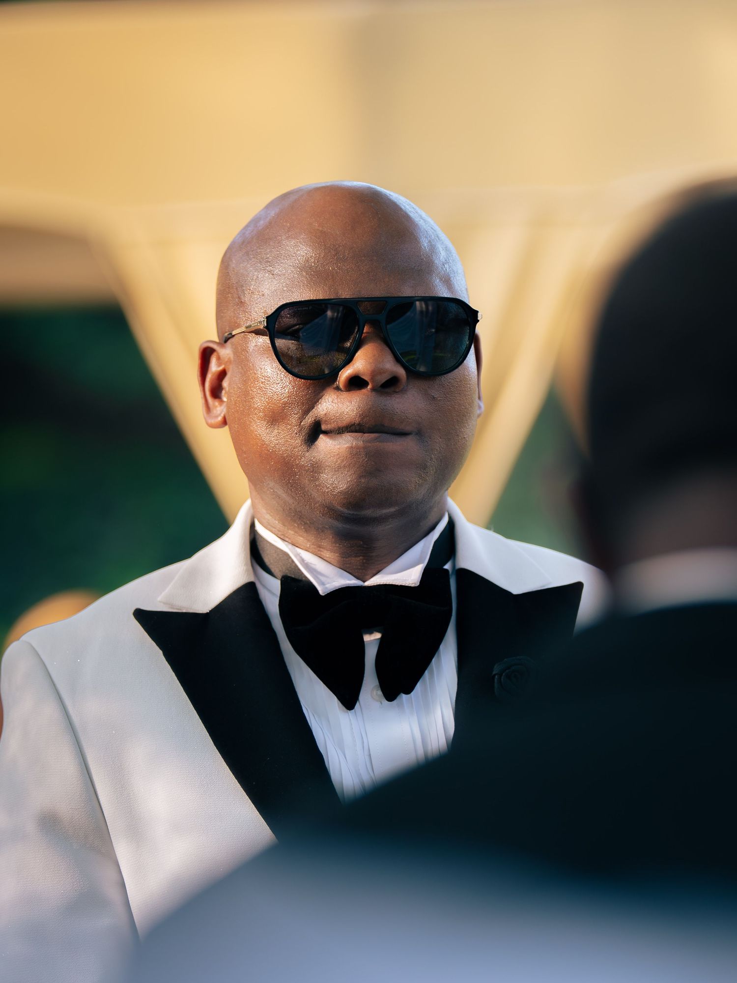 Bald man in white tuxedo jacket with black bow tie and sunglasses at formal event.
