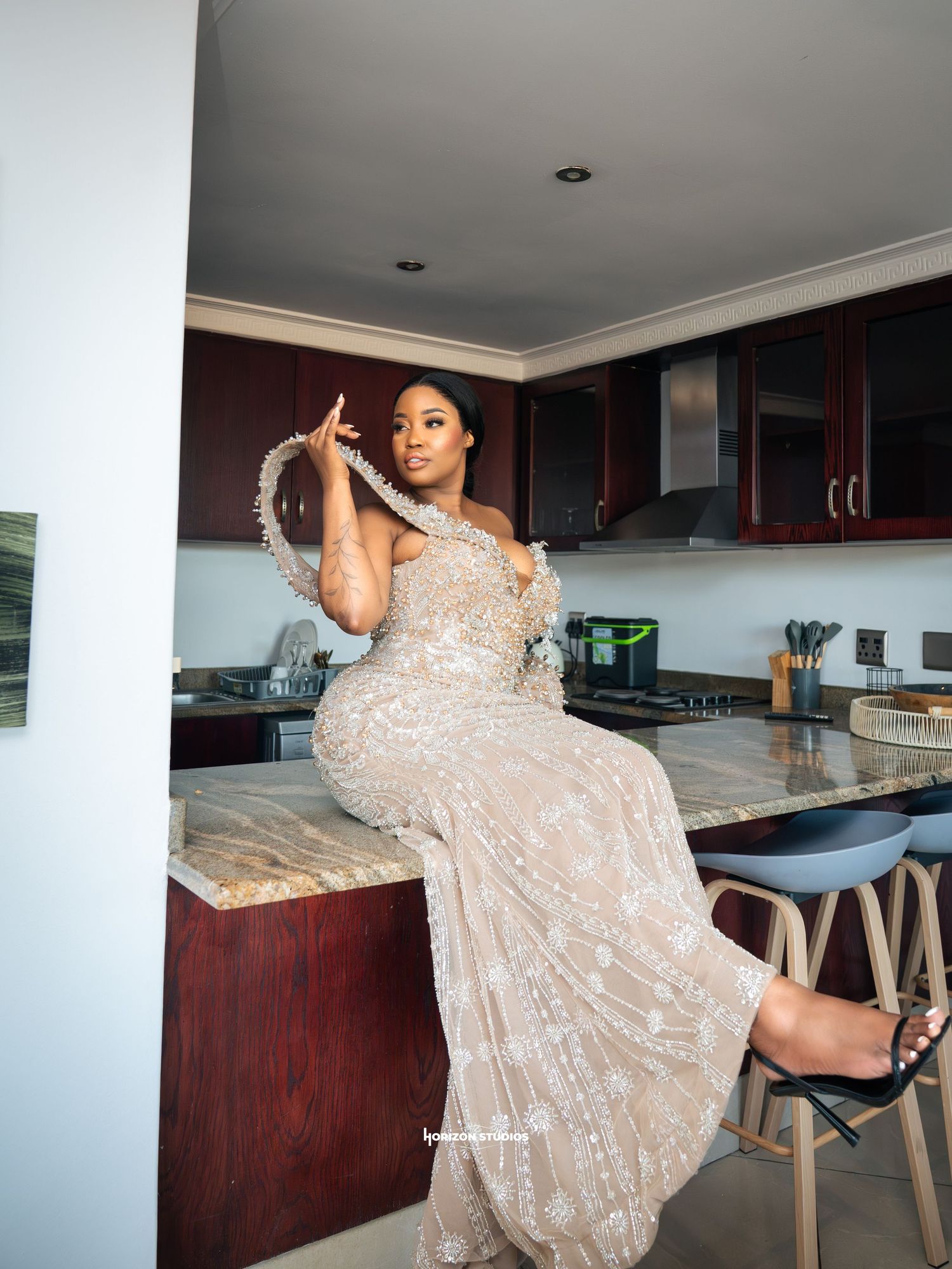 Woman in elegant beaded gown poses on kitchen counter, wearing stunning embellished dress with pearl accessories.