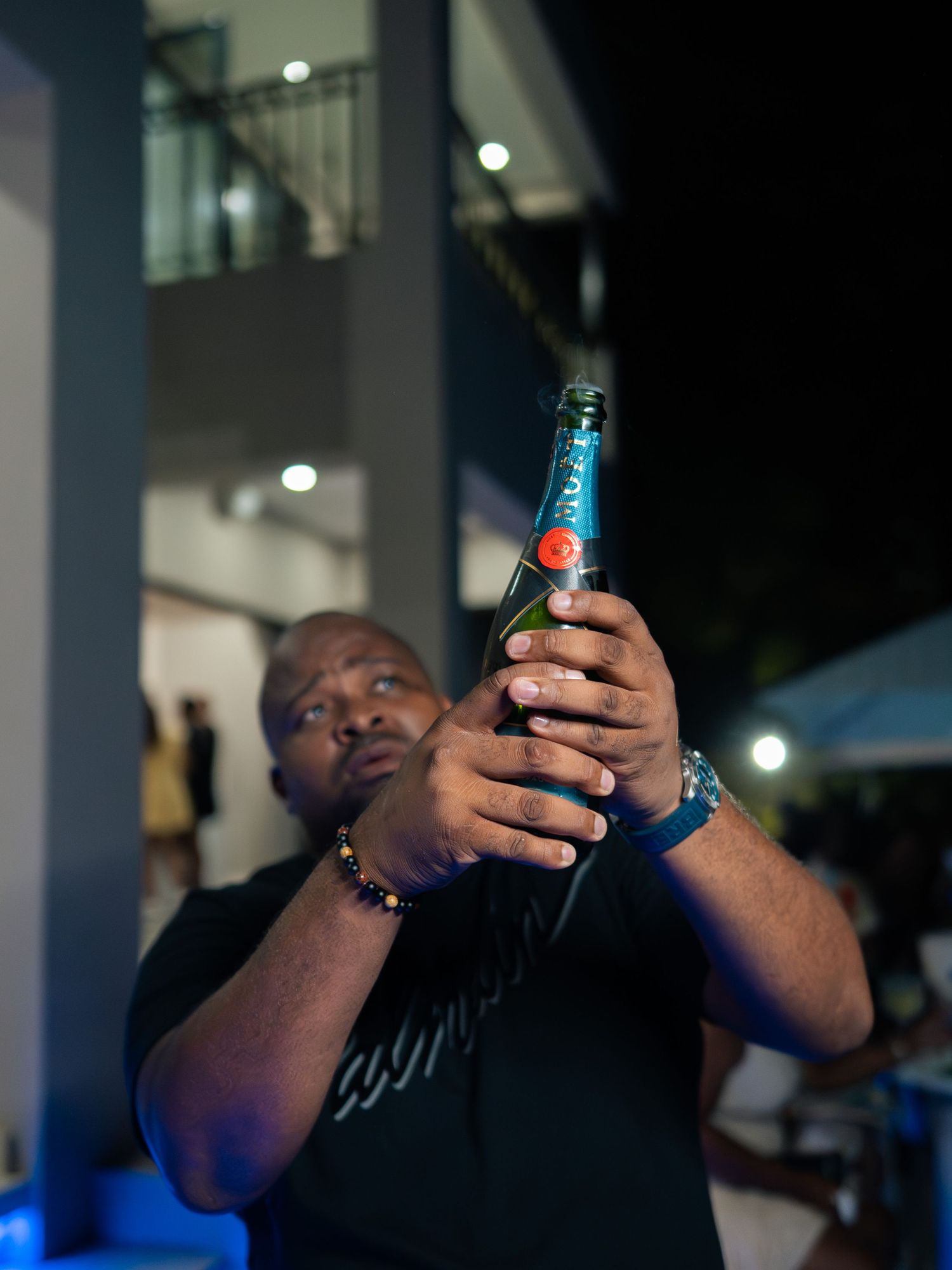 A man holds up a blue beer bottle at an outdoor nighttime party event.