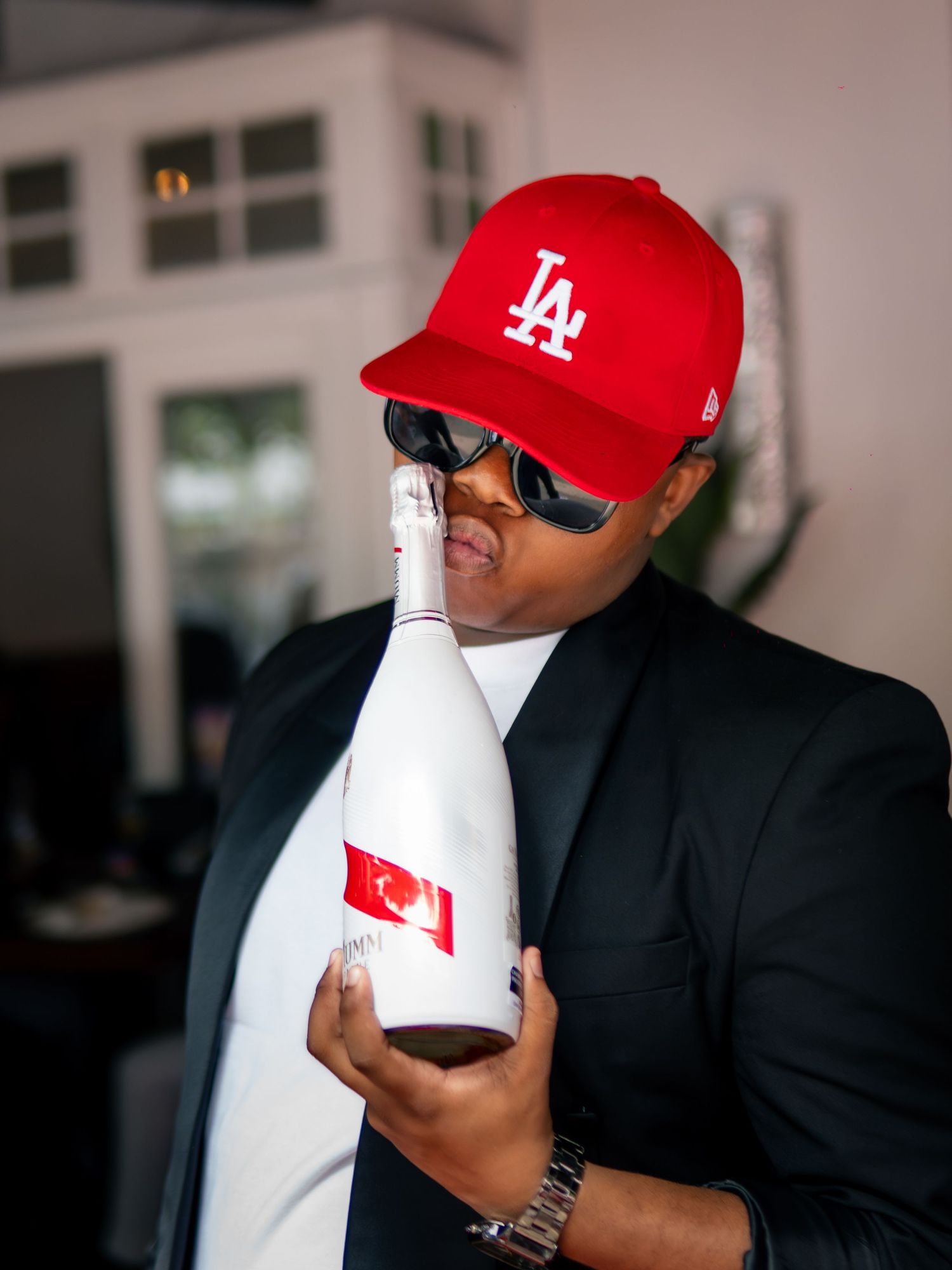 Person wearing red LA cap and sunglasses holding a white bottle at an indoor event.