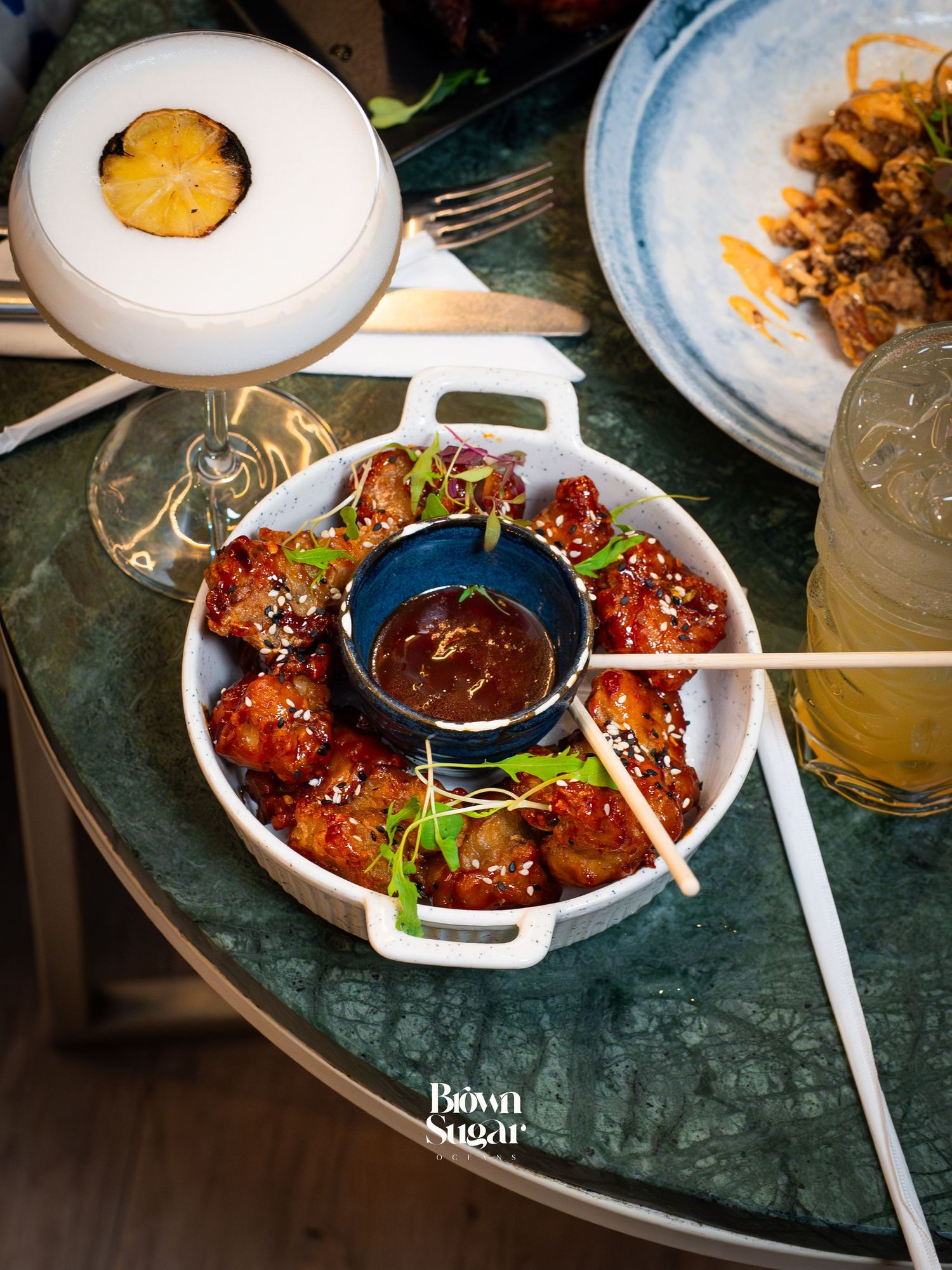 Glazed chicken bites with sesame seeds and dipping sauce in a white bowl, served alongside cocktails at Brown Sugar.