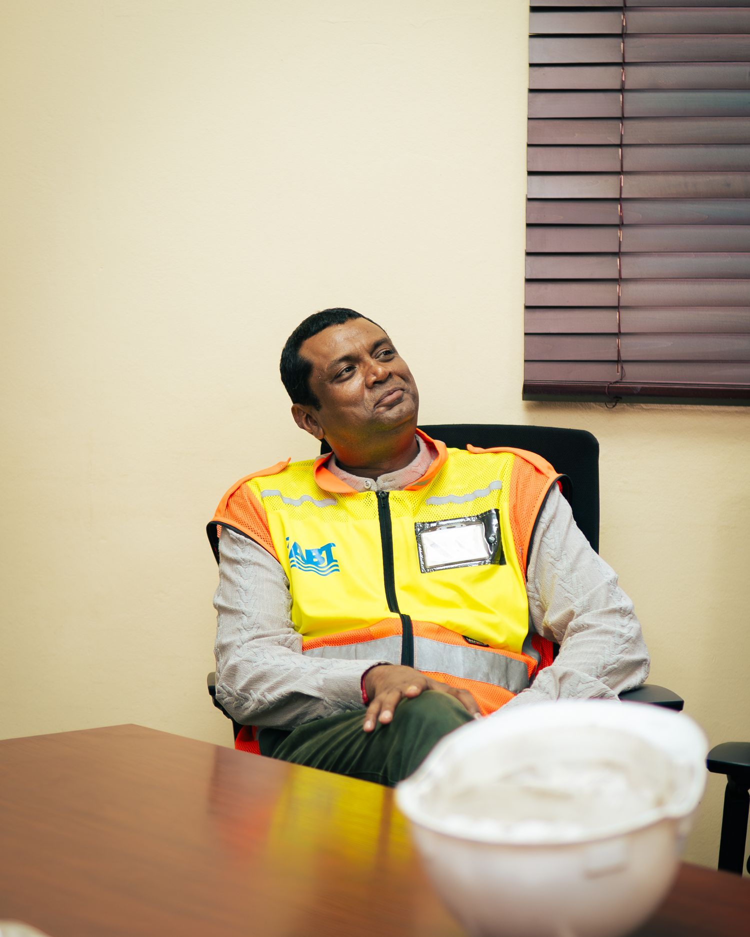 Worker in high-visibility safety vest and hard hat sits at office table, looking thoughtful.