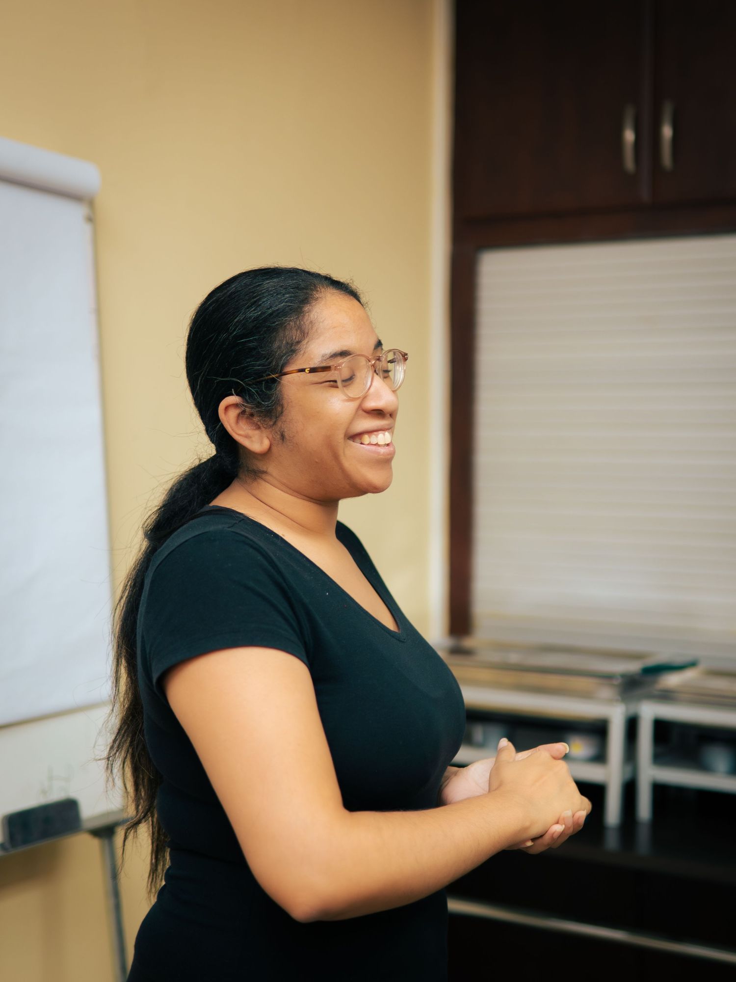 Smiling young woman with glasses and dark hair presenting in a classroom setting, wearing a black top.