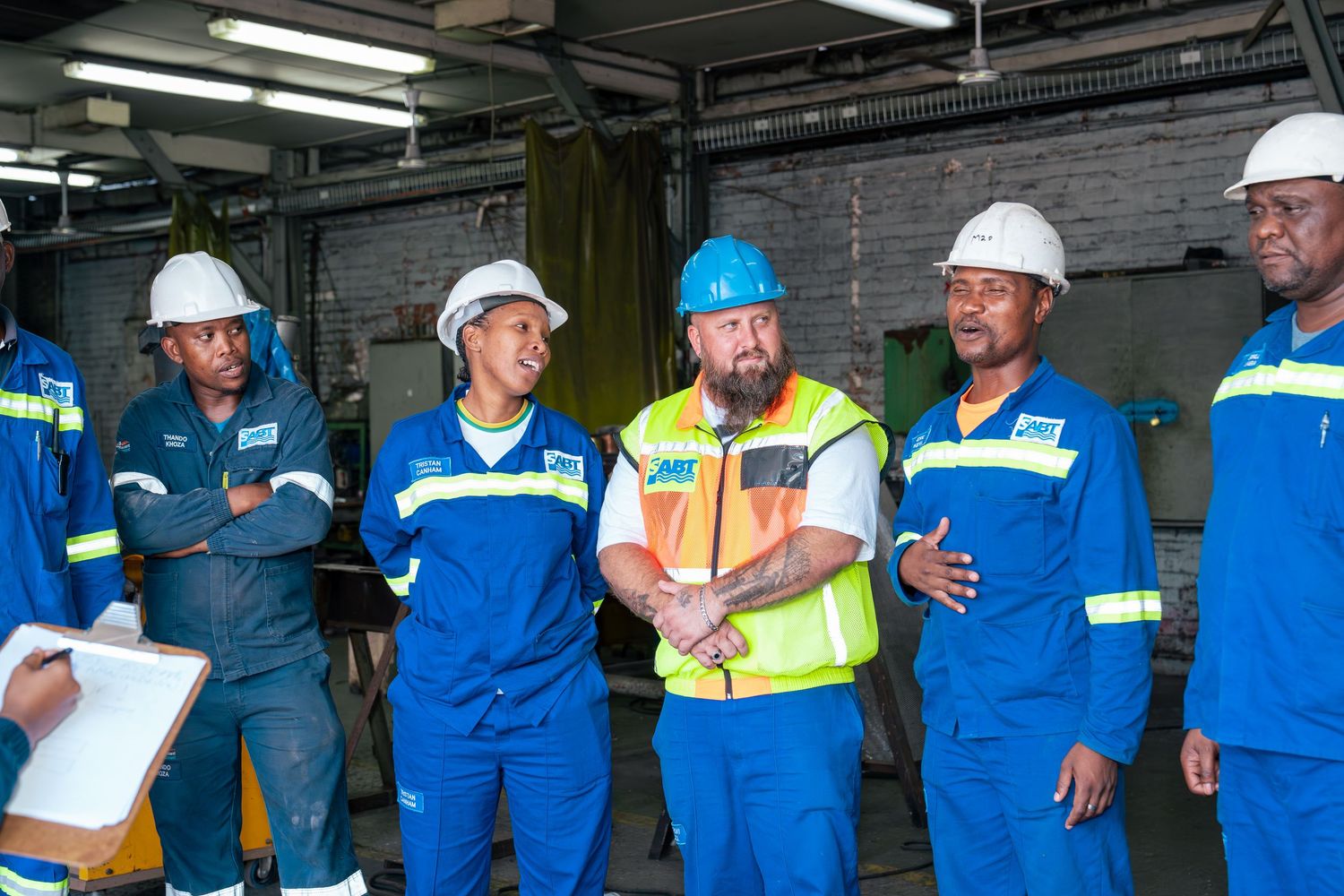 Workers in blue safety uniforms and hard hats collaborate during an industrial workplace briefing led by a supervisor in a yellow vest.