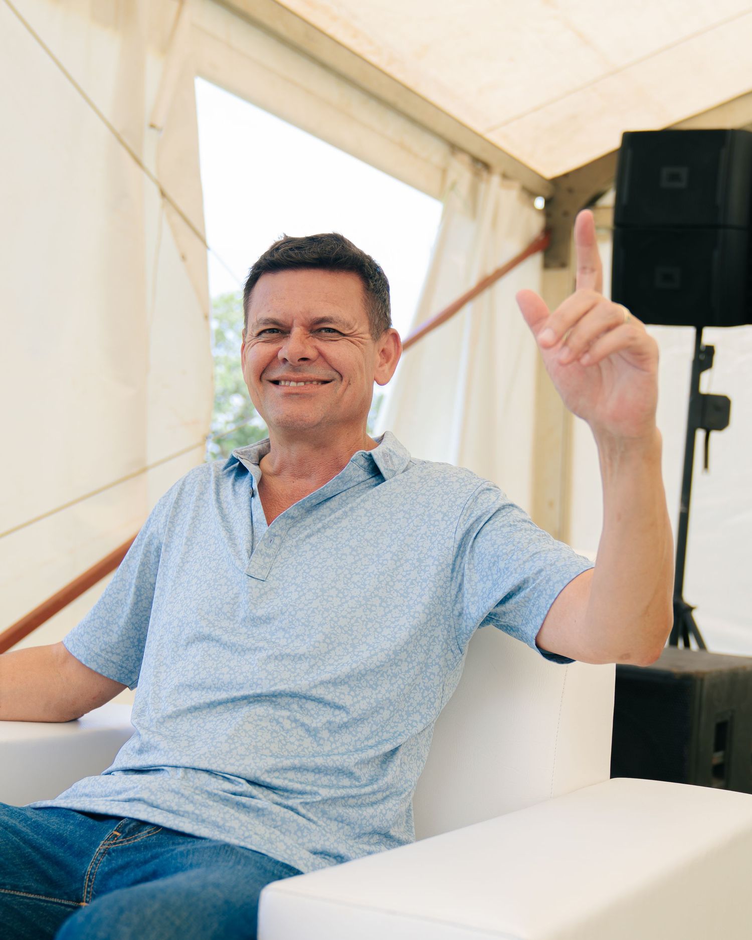 Smiling middle-aged man in light blue polo shirt raising index finger while seated at an outdoor event.
