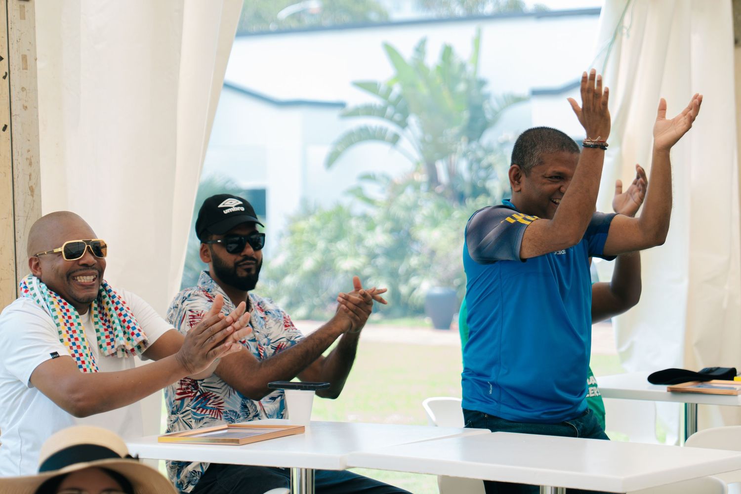 Three men clapping and celebrating outdoors at a sunny event, wearing colorful casual attire.