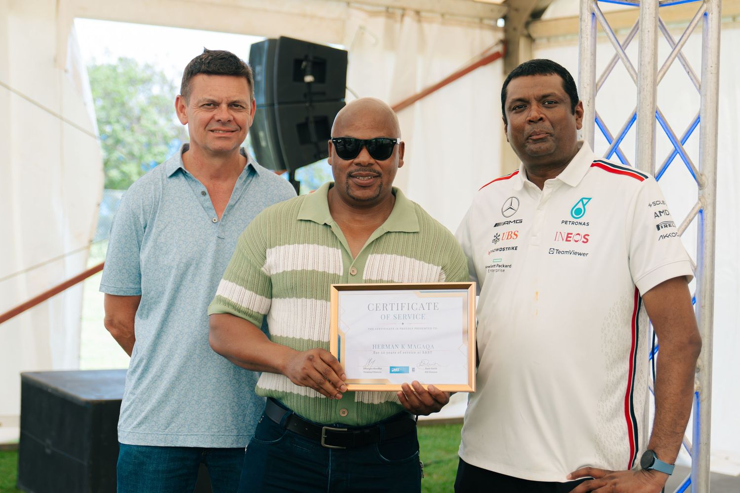 Three men pose outdoors holding a framed certificate of service at a formal recognition ceremony.
