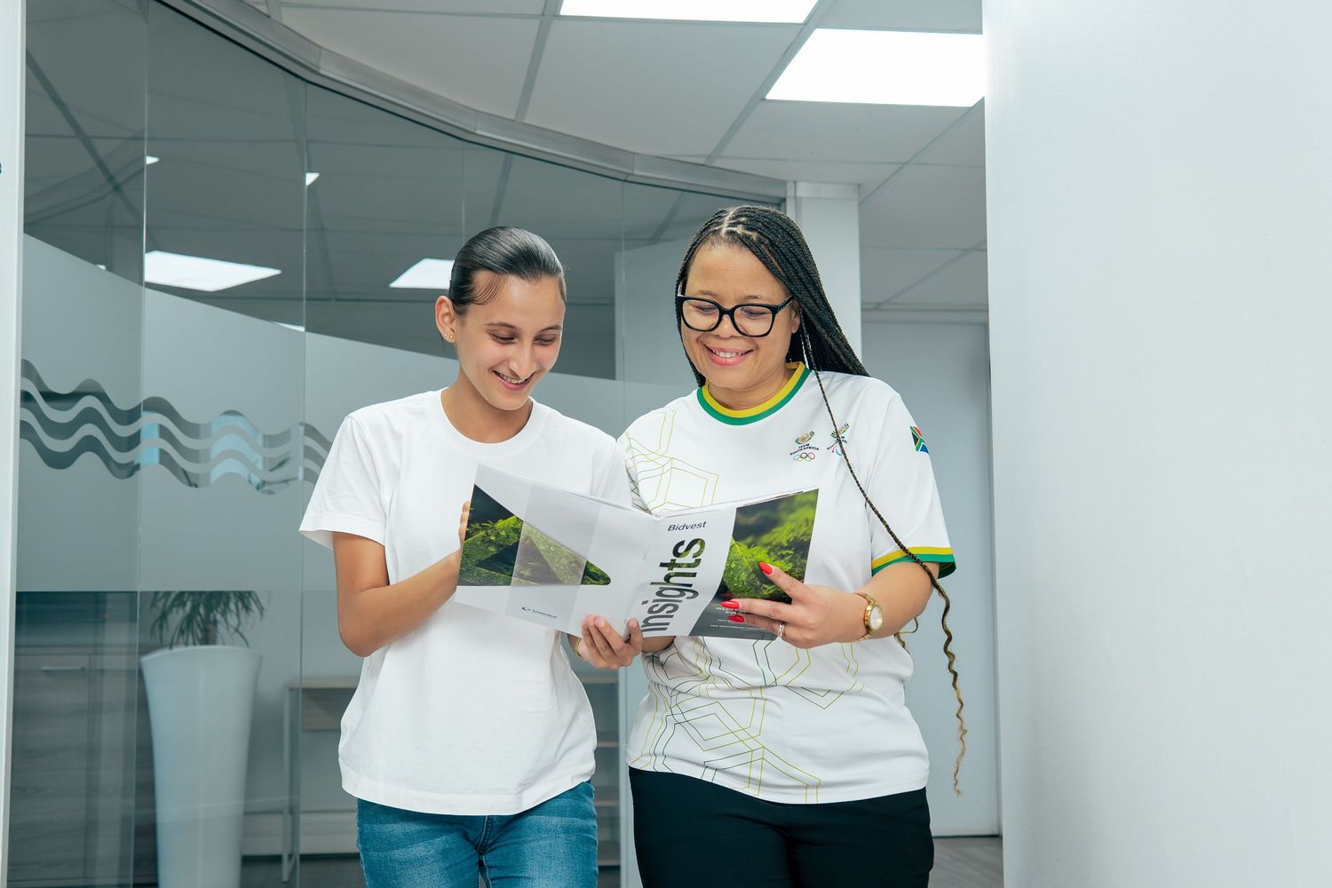 Two women in white t-shirts reviewing a green brochure together in a modern office hallway.