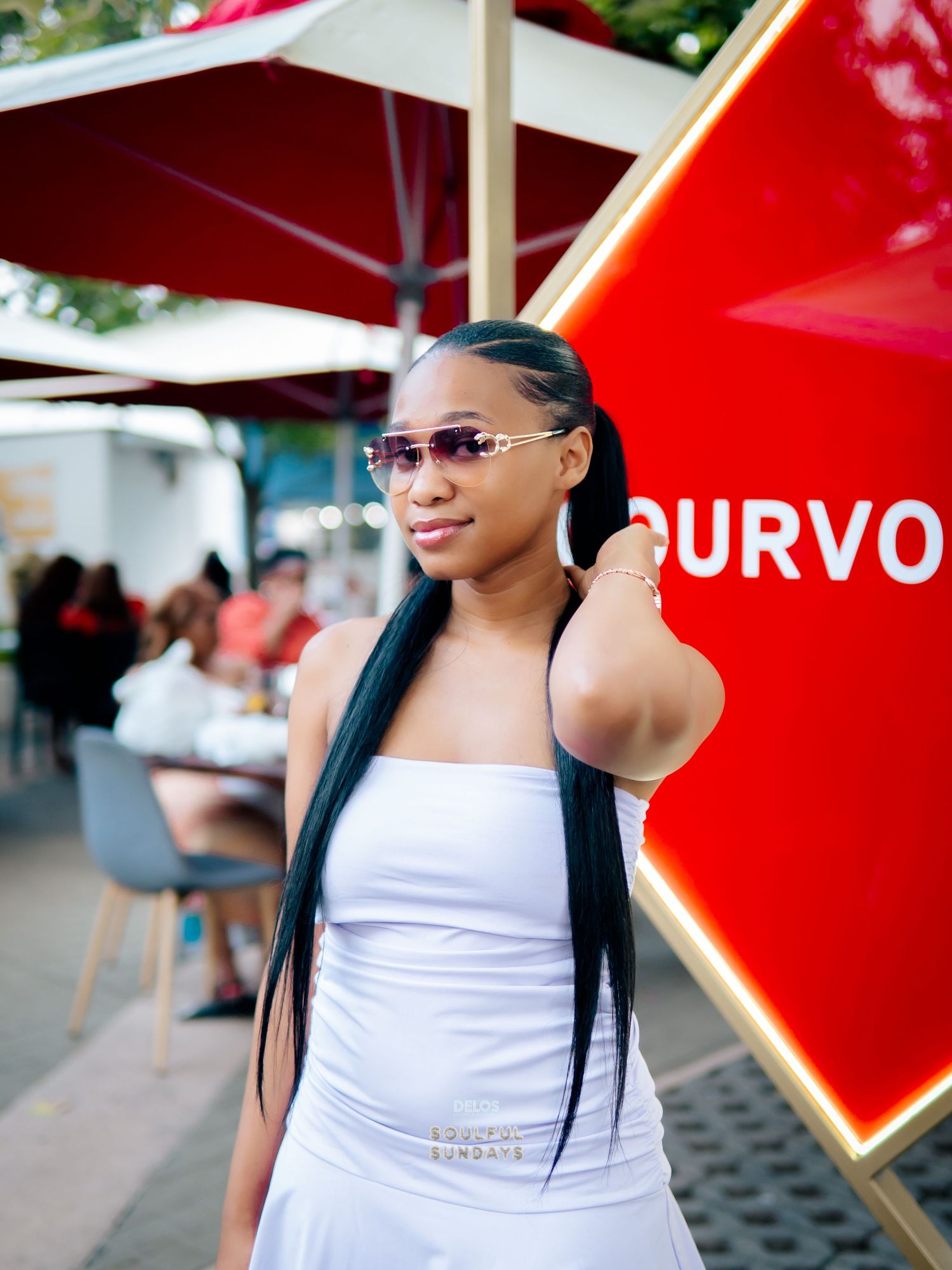Young woman in white dress and rimless sunglasses poses outdoors near a red sign at a casual venue.