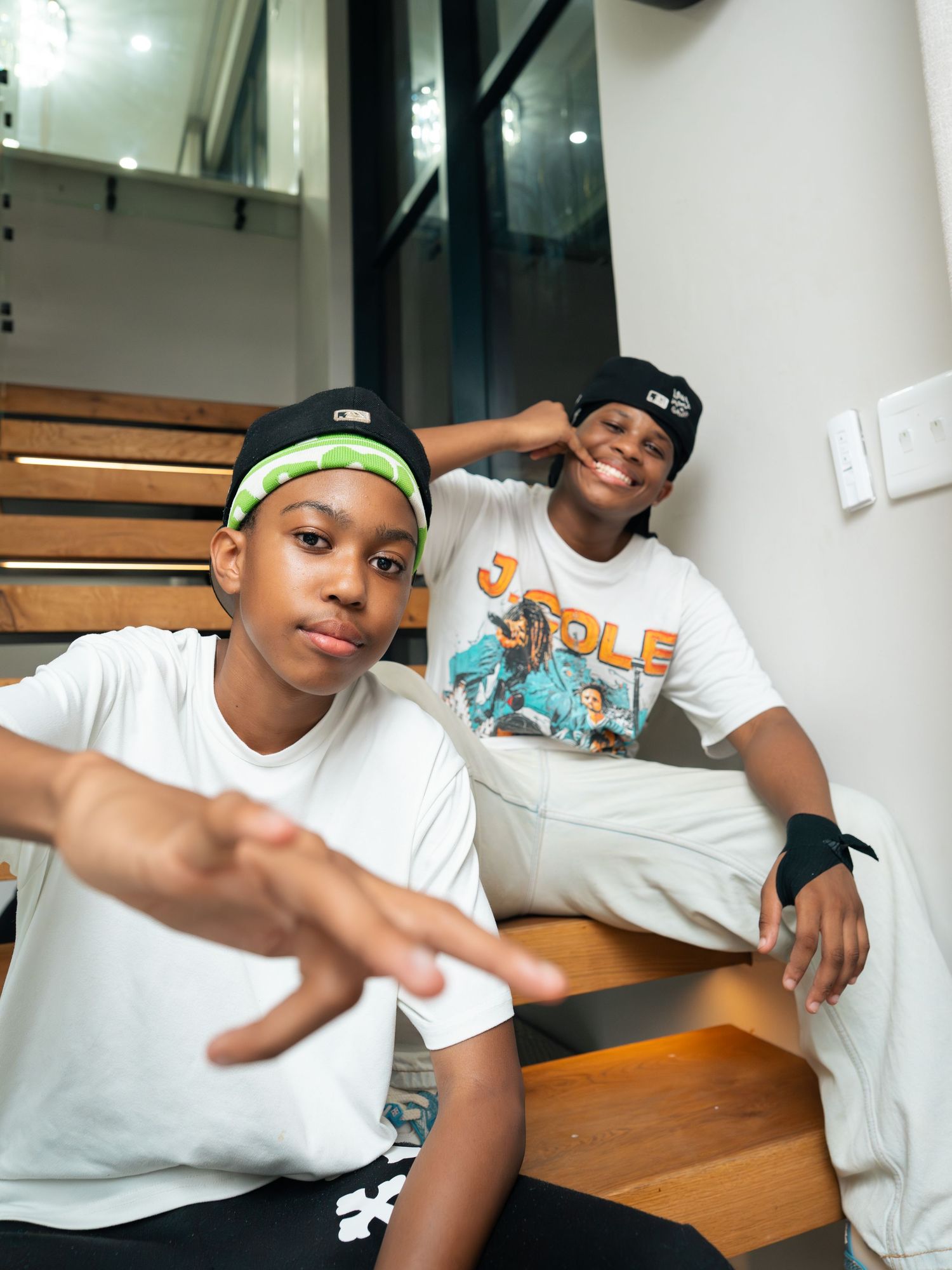 Two young boys in white tees and snapback hats pose on stairs, one reaching toward camera.