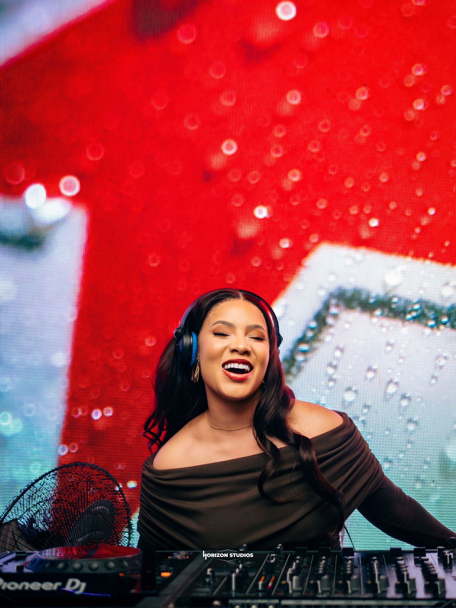 Female DJ smiling behind Pioneer DJ decks, wearing headphones and off-shoulder top, with red backdrop.
