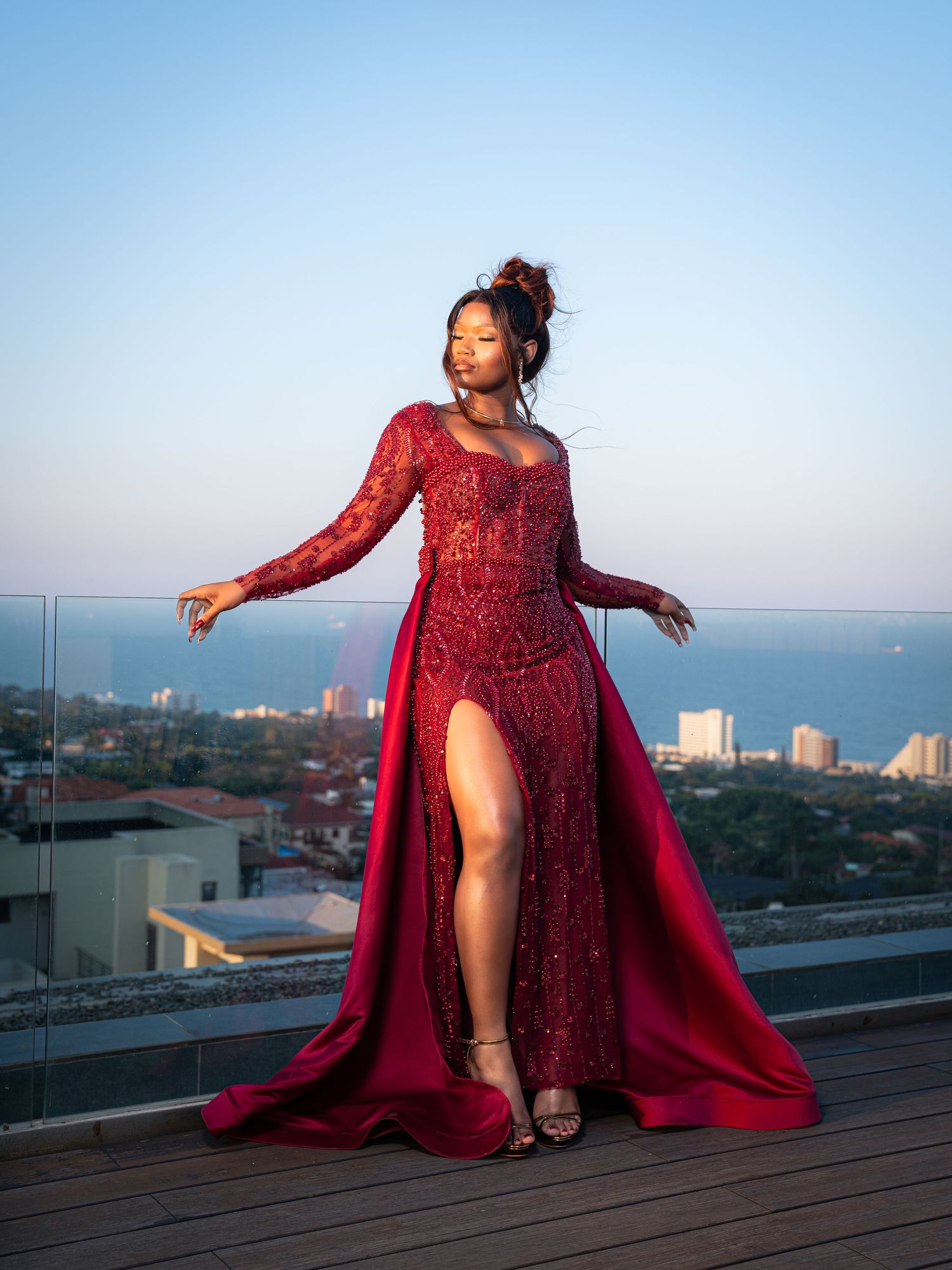 Woman in red beaded gown with dramatic train posing on rooftop terrace with ocean city views.