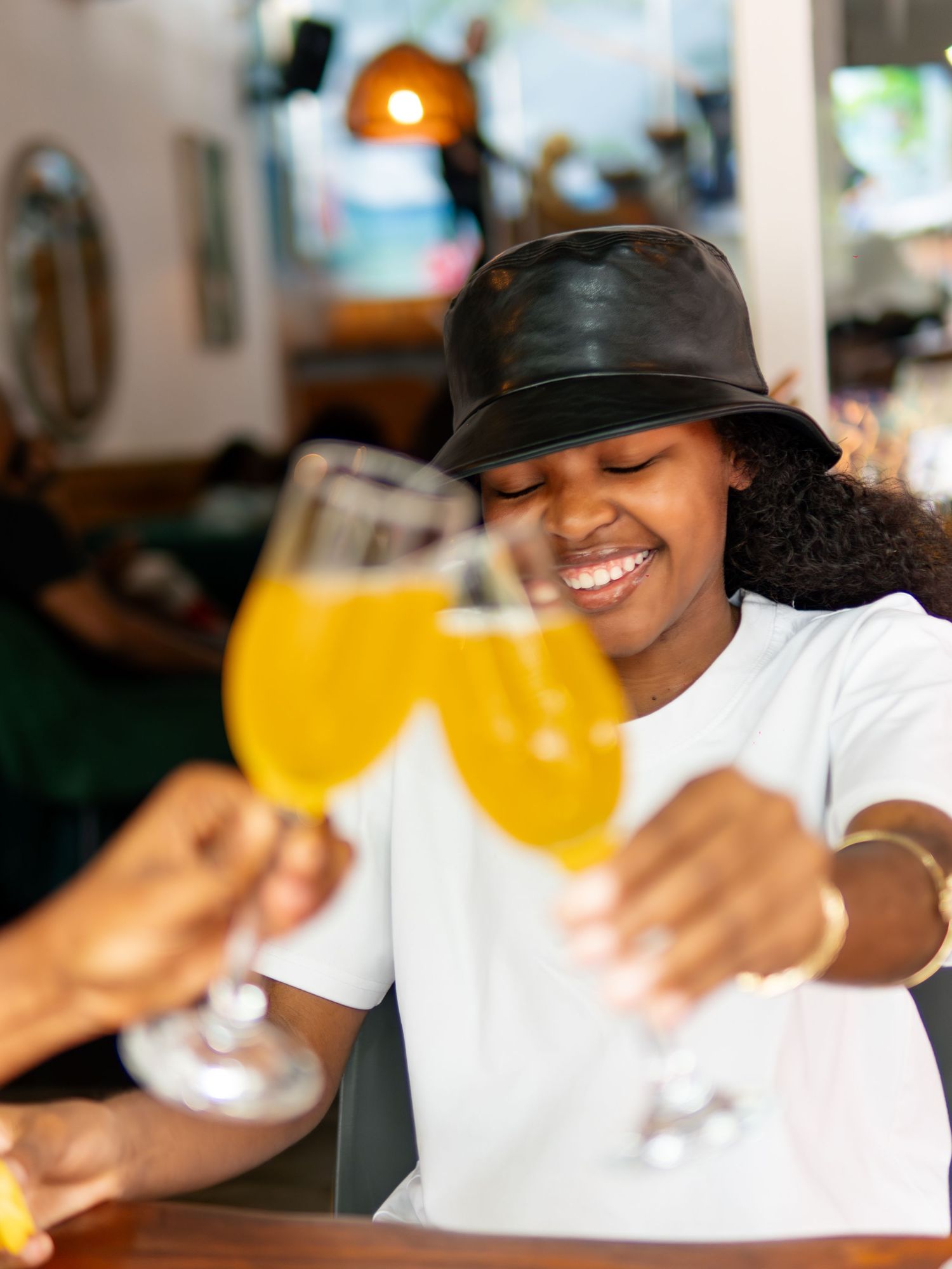Smiling young woman in black bucket hat toasting with orange mimosas at a bar with friends.