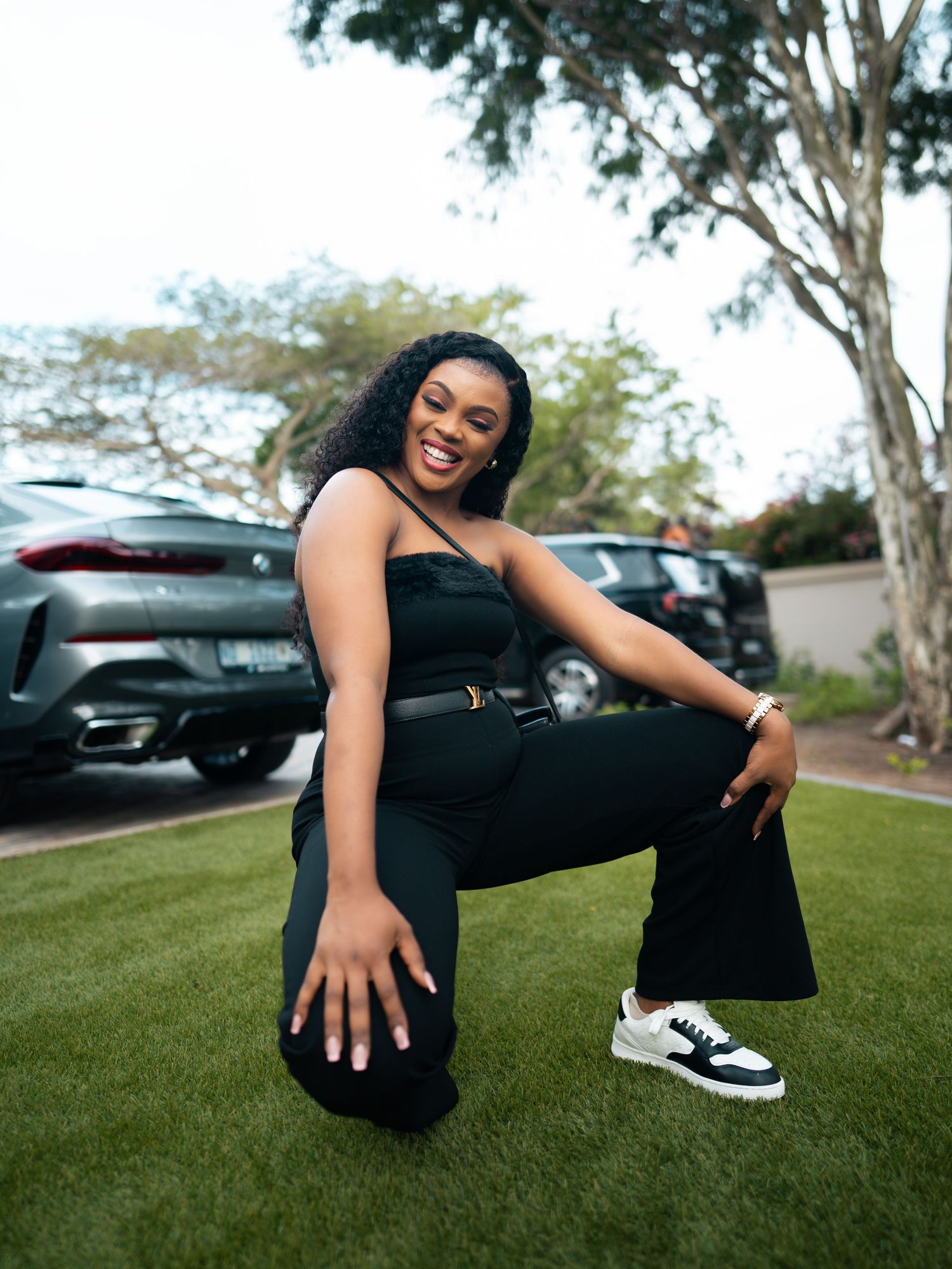 A young Black woman in a black jumpsuit and sneakers poses crouching on green grass near parked cars and a tree.
