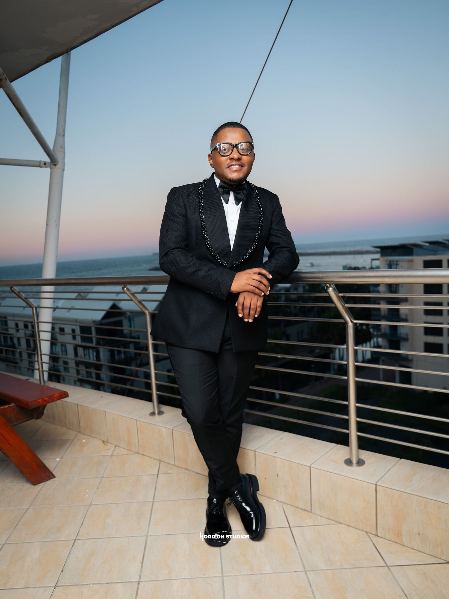 Elegant man in black tuxedo posing on rooftop terrace at sunset with ocean view backdrop.