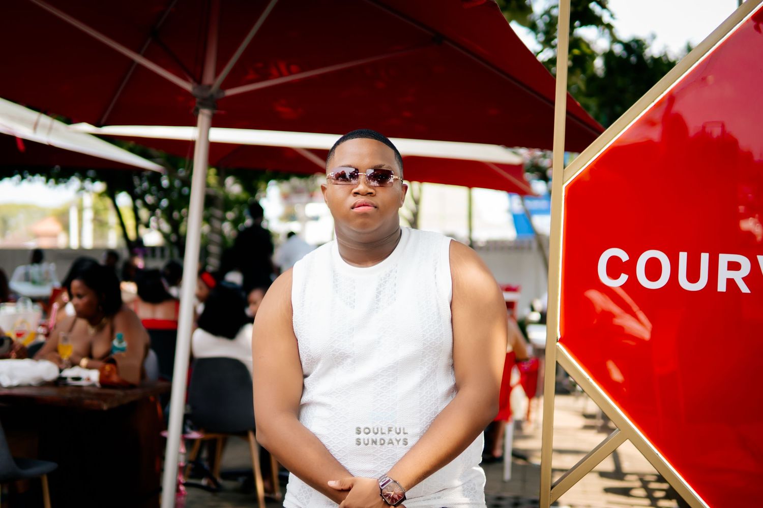 Young man in white sleeveless shirt and sunglasses posing outdoors at a vibrant social event near a red sign.