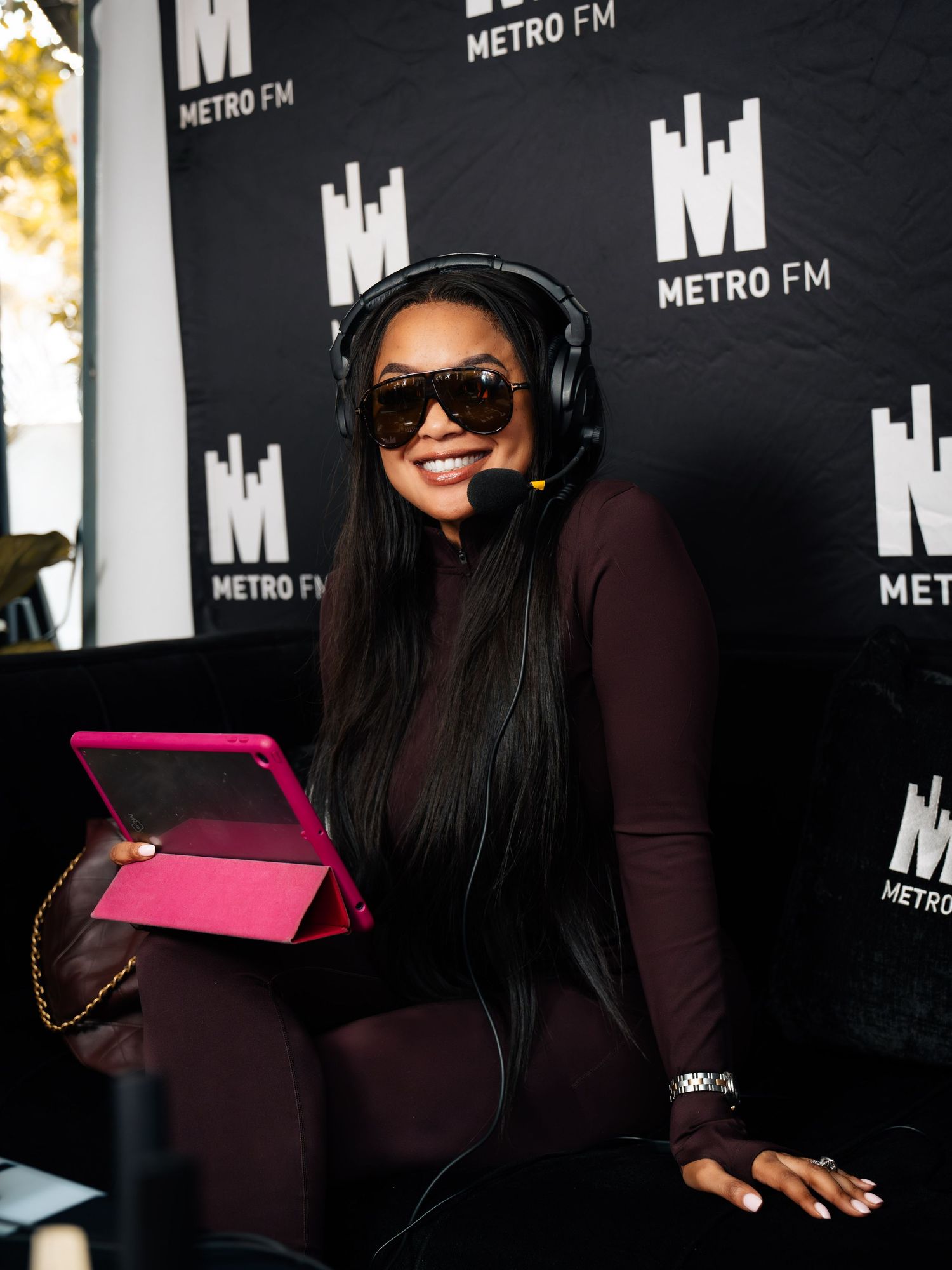 Woman DJ with headphones and sunglasses holds pink tablet at Metro FM radio broadcast event.