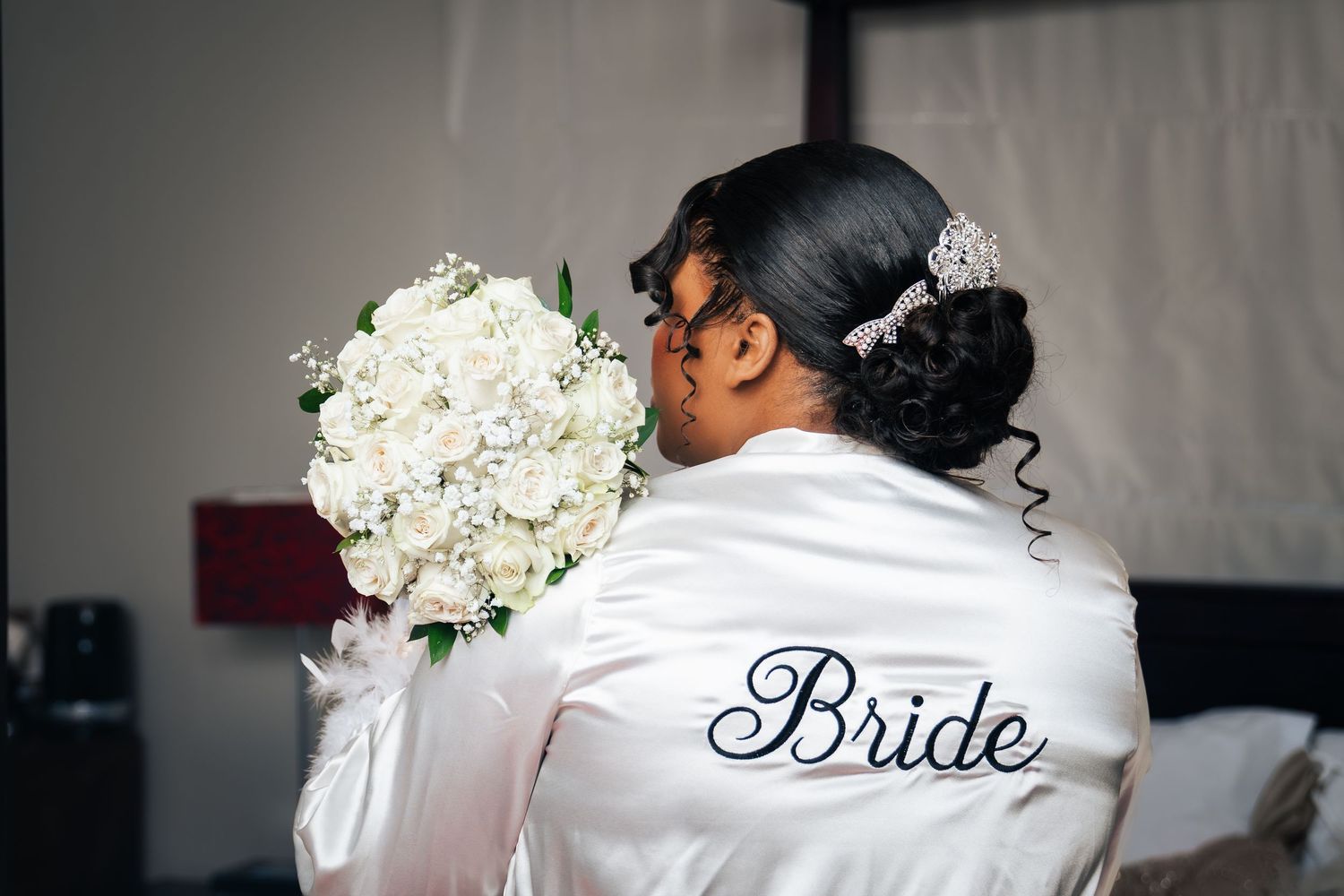 Bride in white satin robe holding elegant white rose bouquet, viewed from behind on wedding day.