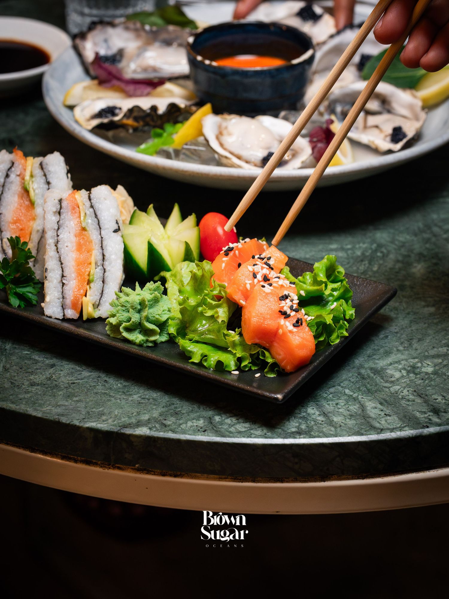 Chopsticks picking up fresh salmon sushi on a dark plate with cucumber and greens at Brown Sugar restaurant.