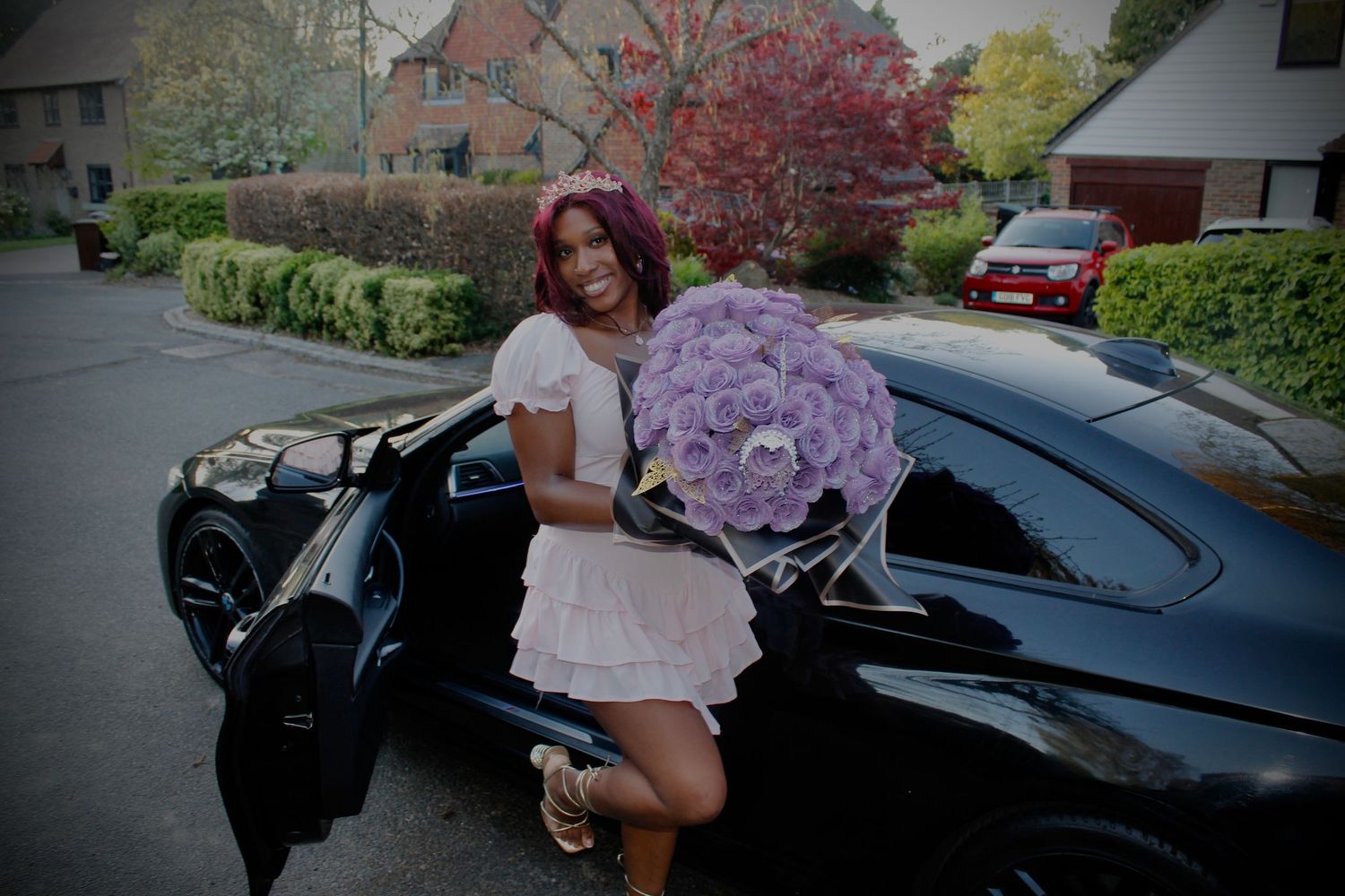 Young woman in white dress holding large purple flower bouquet beside a black luxury car.