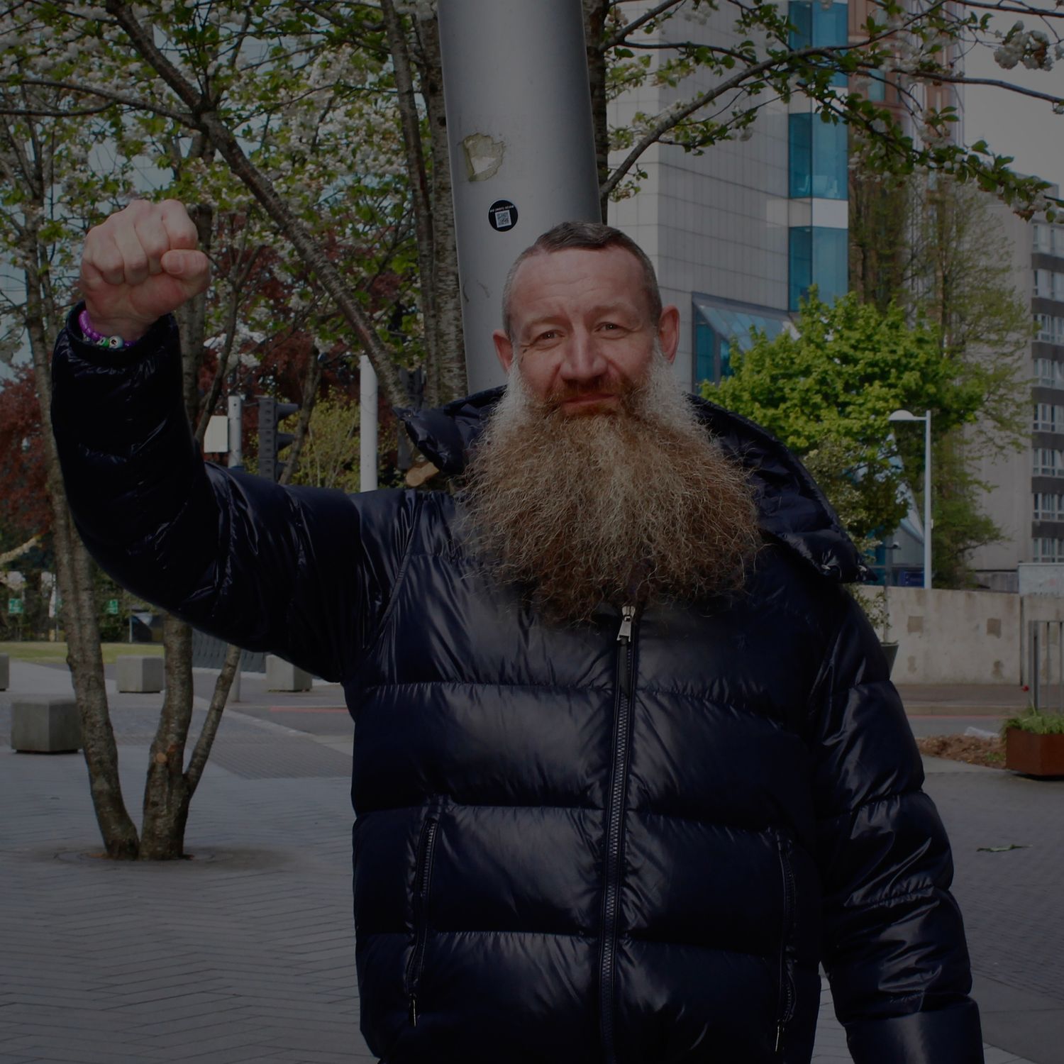 Older man with large grey beard wearing black puffer jacket raising fist outdoors in urban setting.