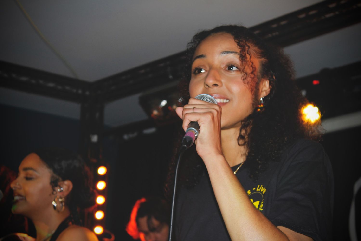 Young woman singing into a microphone on stage at a live music event under warm stage lighting.
