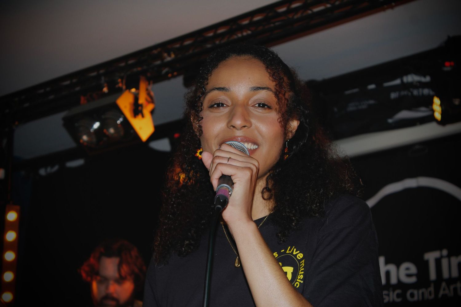 Young woman smiling and singing into a microphone on stage at the Tin Music and Arts venue.