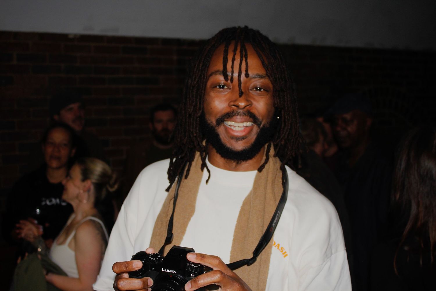 Smiling man with dreadlocks holding a camera at an indoor event wearing a white t-shirt.