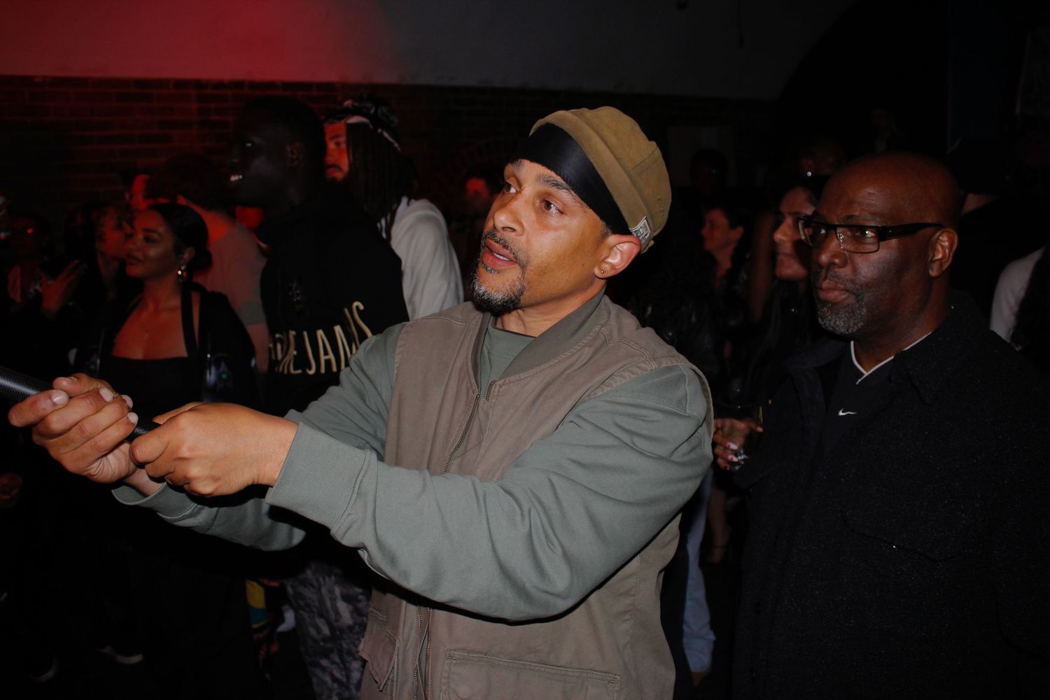 Man pointing while holding a phone at an indoor music event with red ambient lighting.