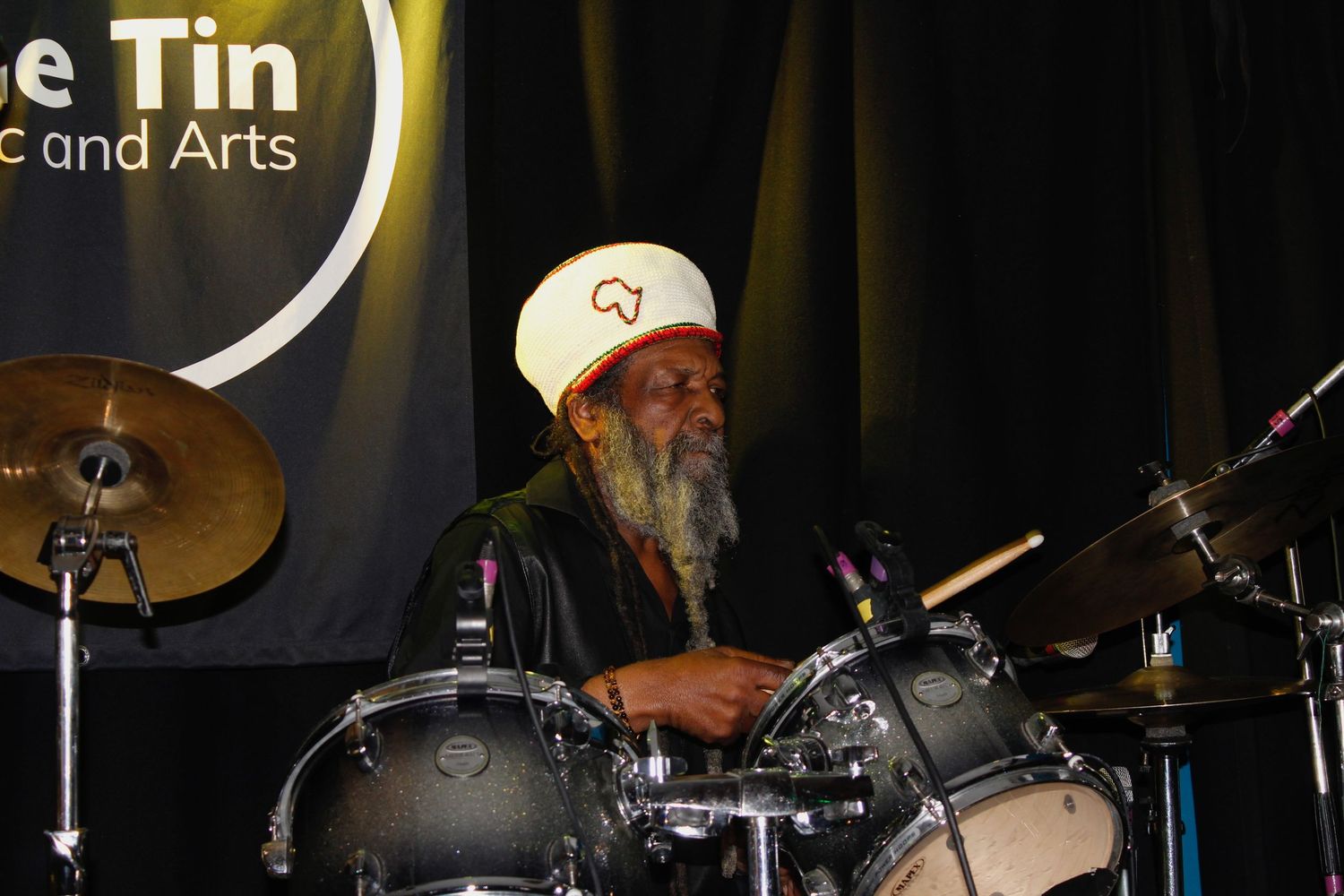 Elderly musician with white beard and hat playing drums at the Tin Music and Arts venue.