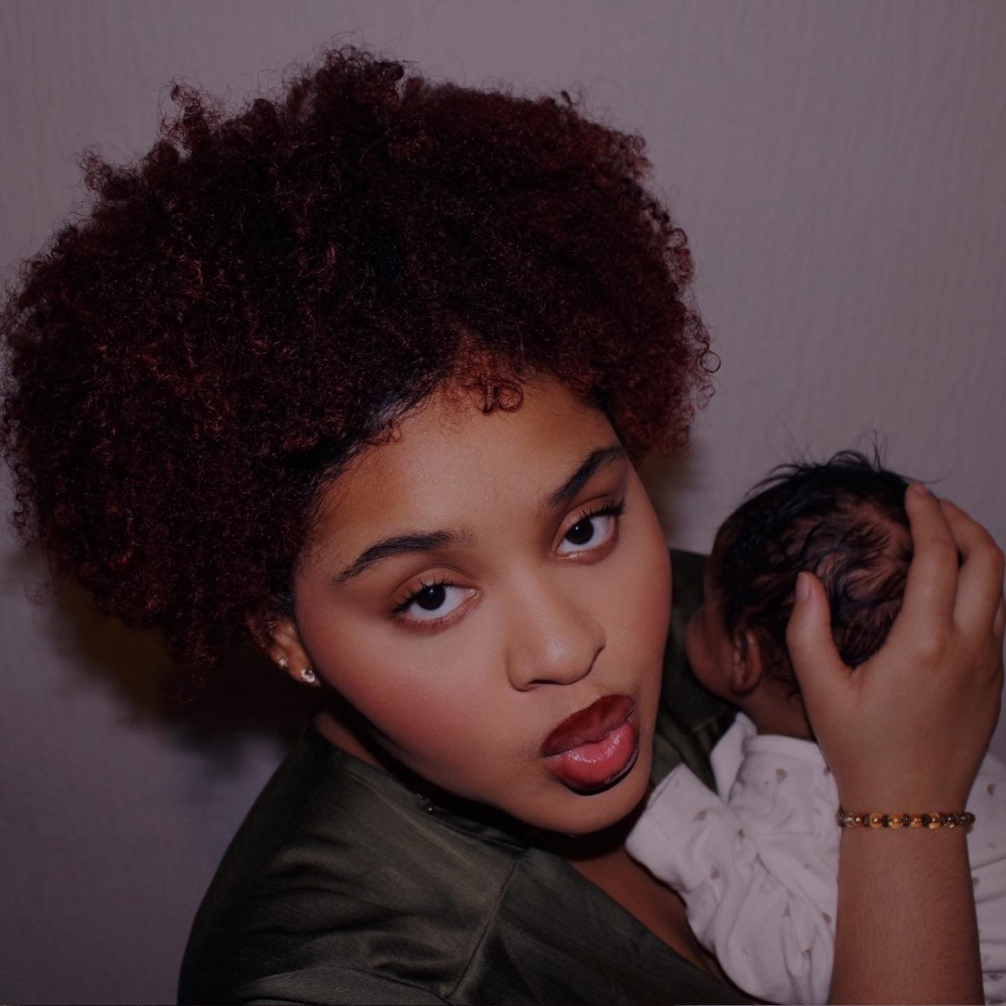 Young woman with dark red curly afro hair holding a small object against a light background.