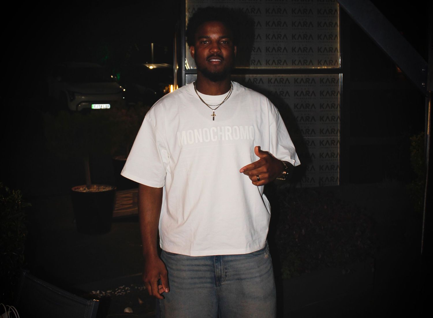 Young man in white graphic t-shirt and jeans posing outdoors at night, pointing finger gesture.