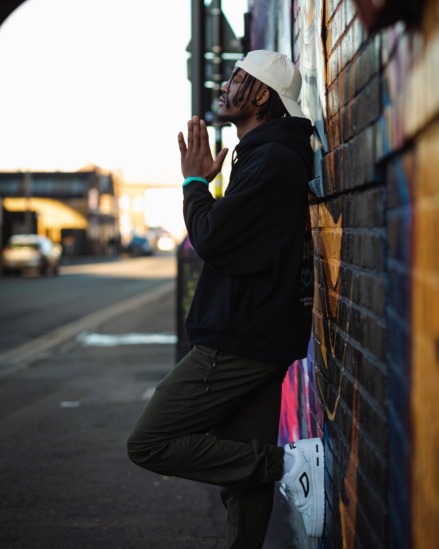 Man in black hoodie posing with hands together against a graffiti-covered brick wall.