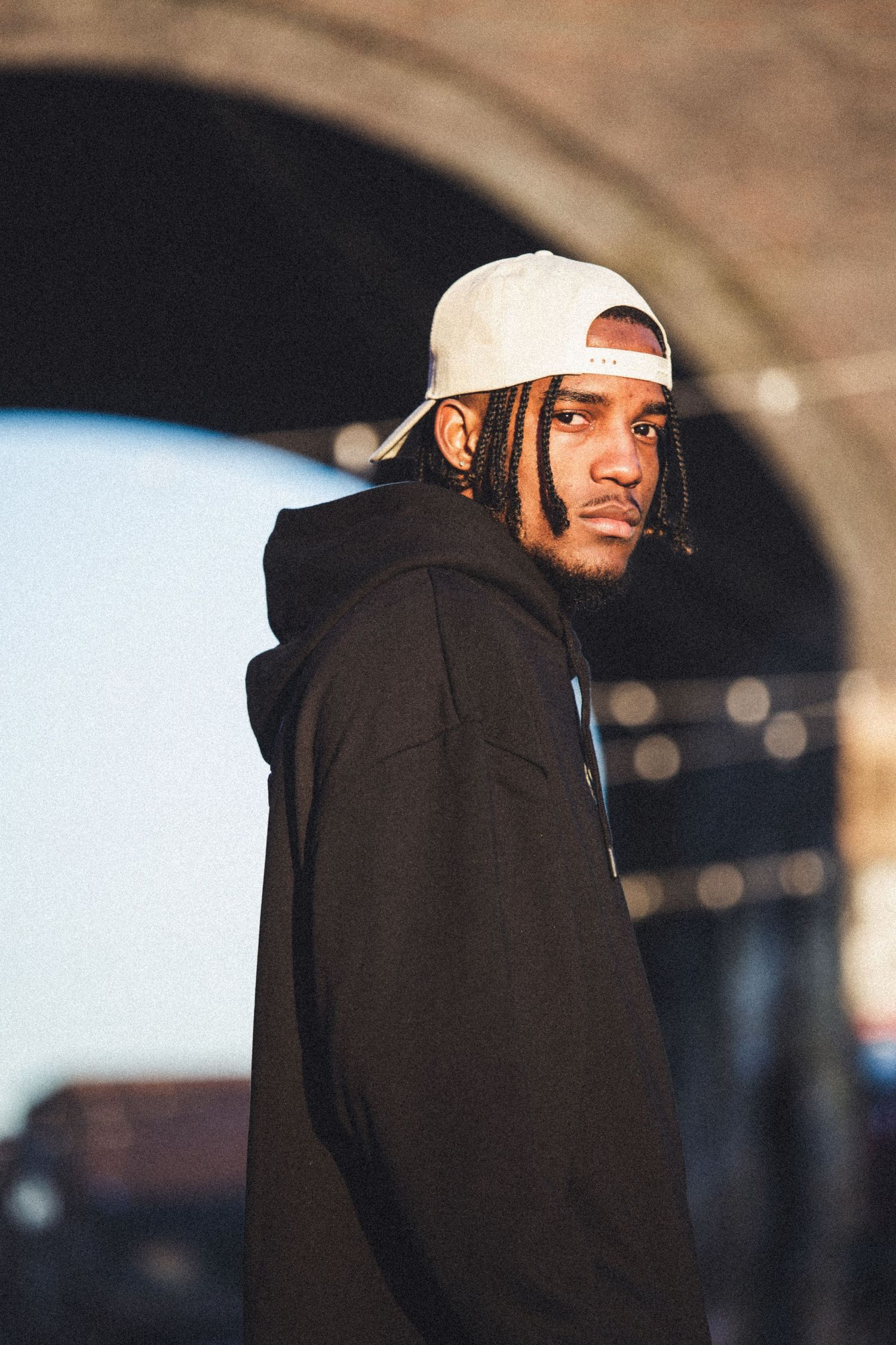 Young man in black hoodie and white snapback cap posing under an arch outdoors.