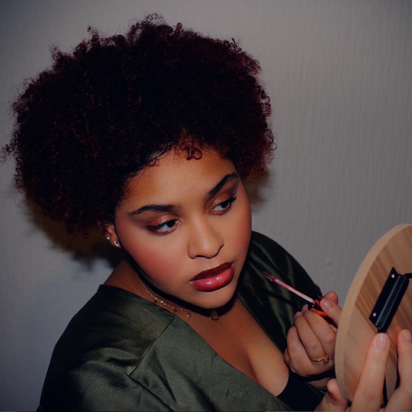 Young woman with natural afro hair applying lip gloss using a handheld mirror in soft lighting.