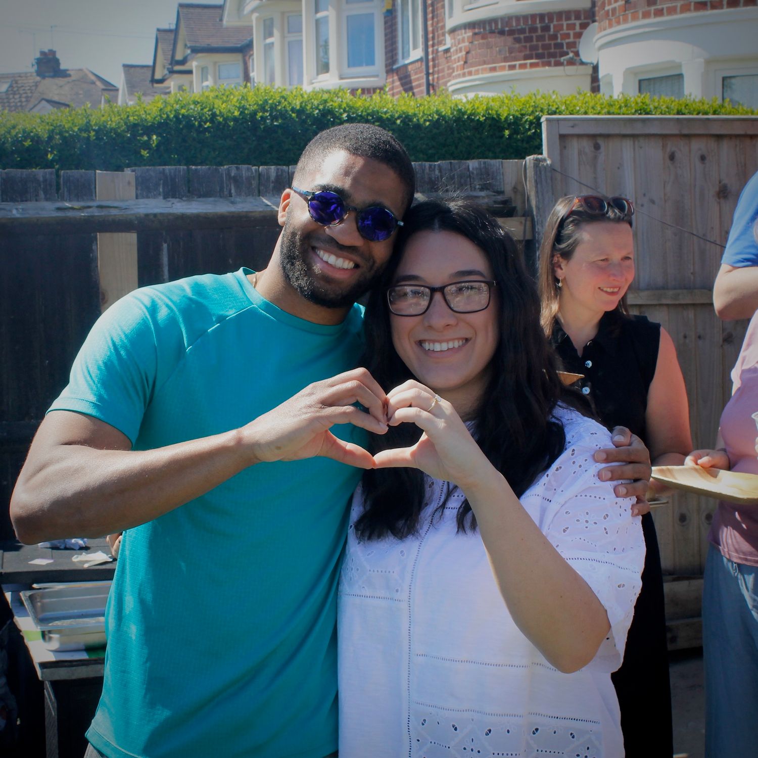Smiling couple making heart shape with hands at outdoor garden party on sunny day.