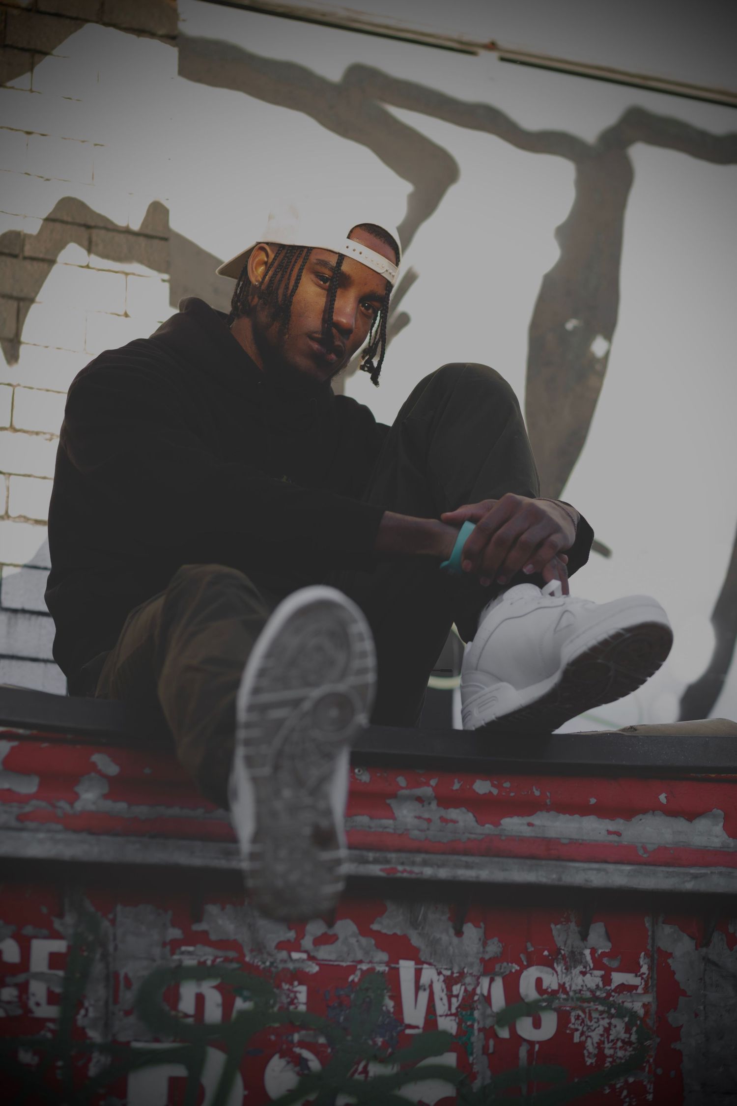Man in black hoodie and white cap sitting on a ledge against a graffiti wall outdoors.