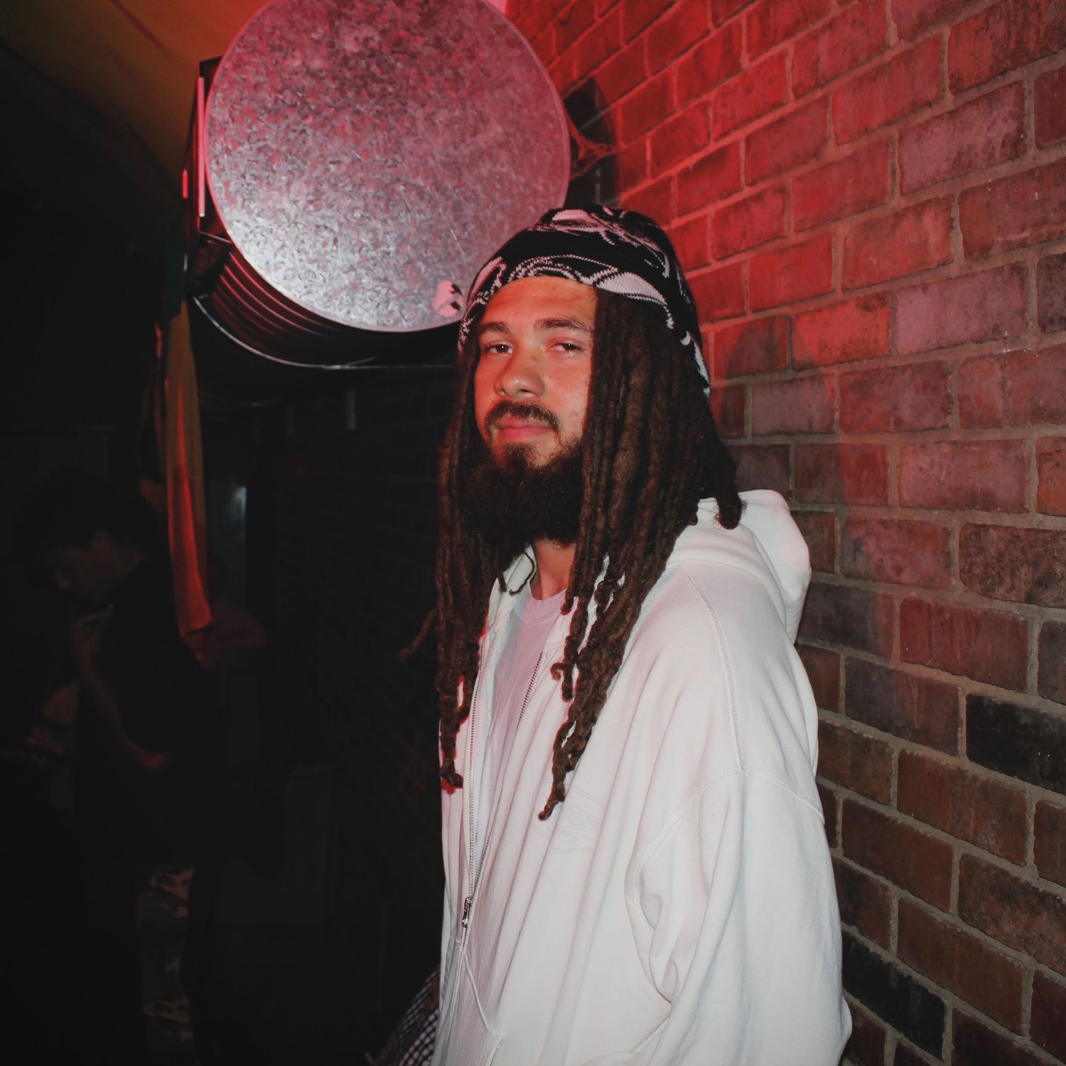 Man with long dreadlocks and bandana wearing a white hoodie standing by a brick wall at a music event.