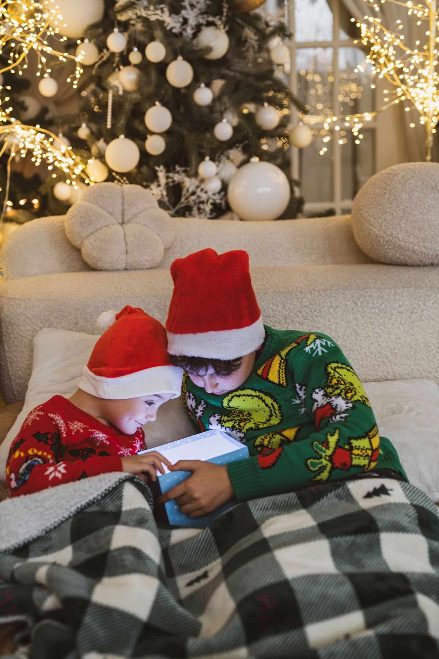 Two children in Santa hats and holiday pajamas lying under blanket using tablet by Christmas tree.