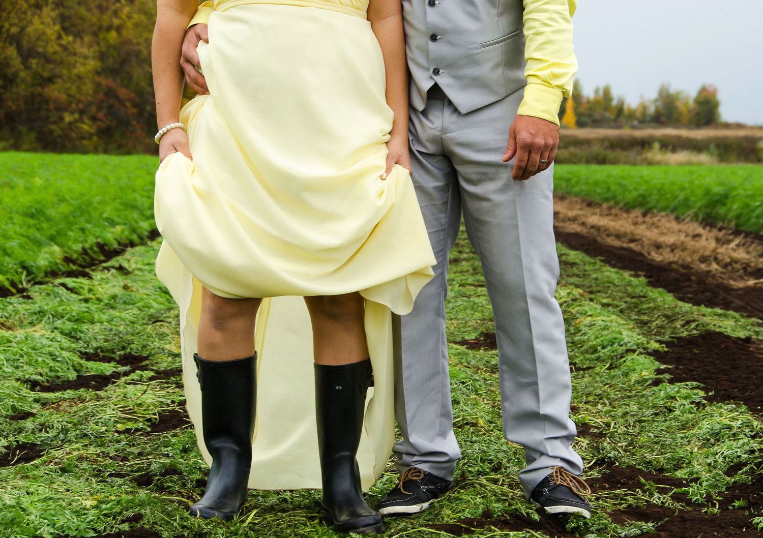 Bride in yellow dress wearing rain boots standing with groom in gray suit in a green farm field.