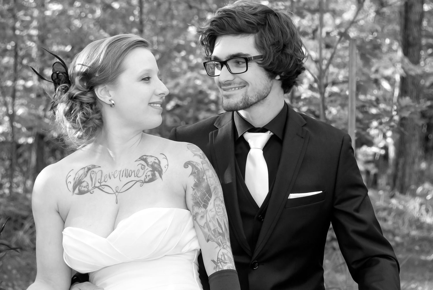 Black and white photo of smiling bride with tattoos and groom in suit sharing a loving look.