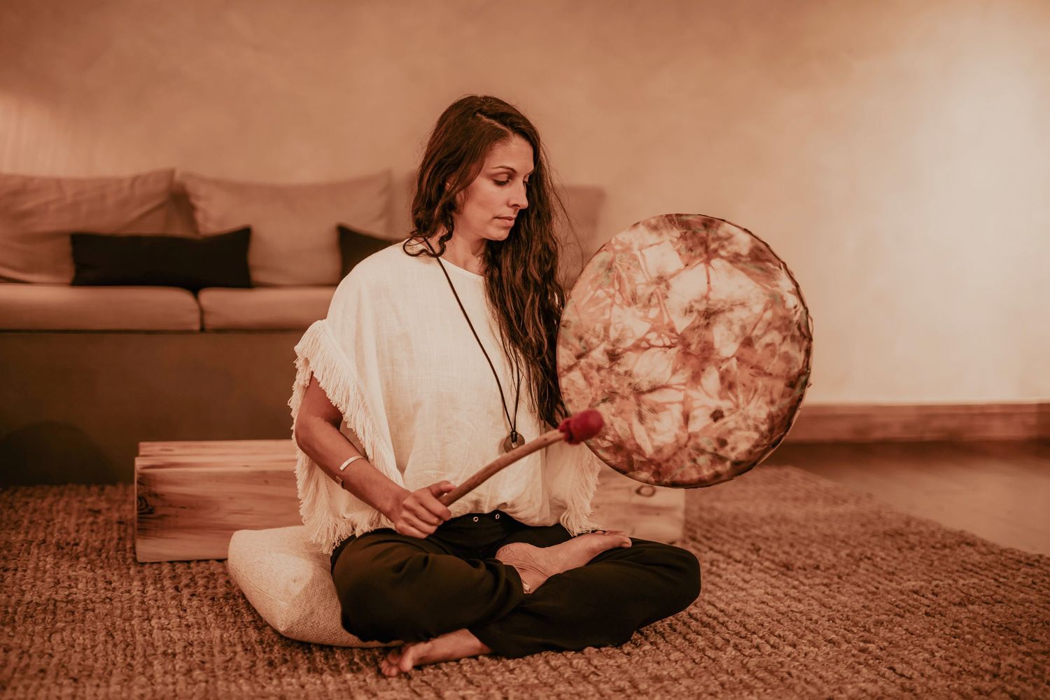 Woman in white poncho sits cross-legged playing a shamanic drum in warm-lit room.