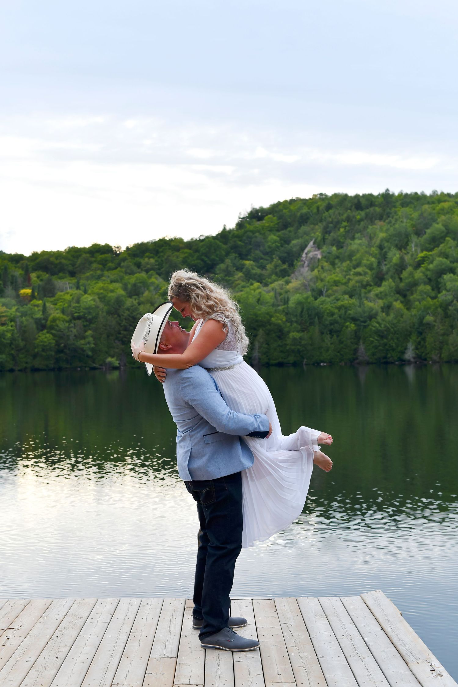 Groom lifting bride in white dress on a wooden dock with a scenic lake and green hills behind them.