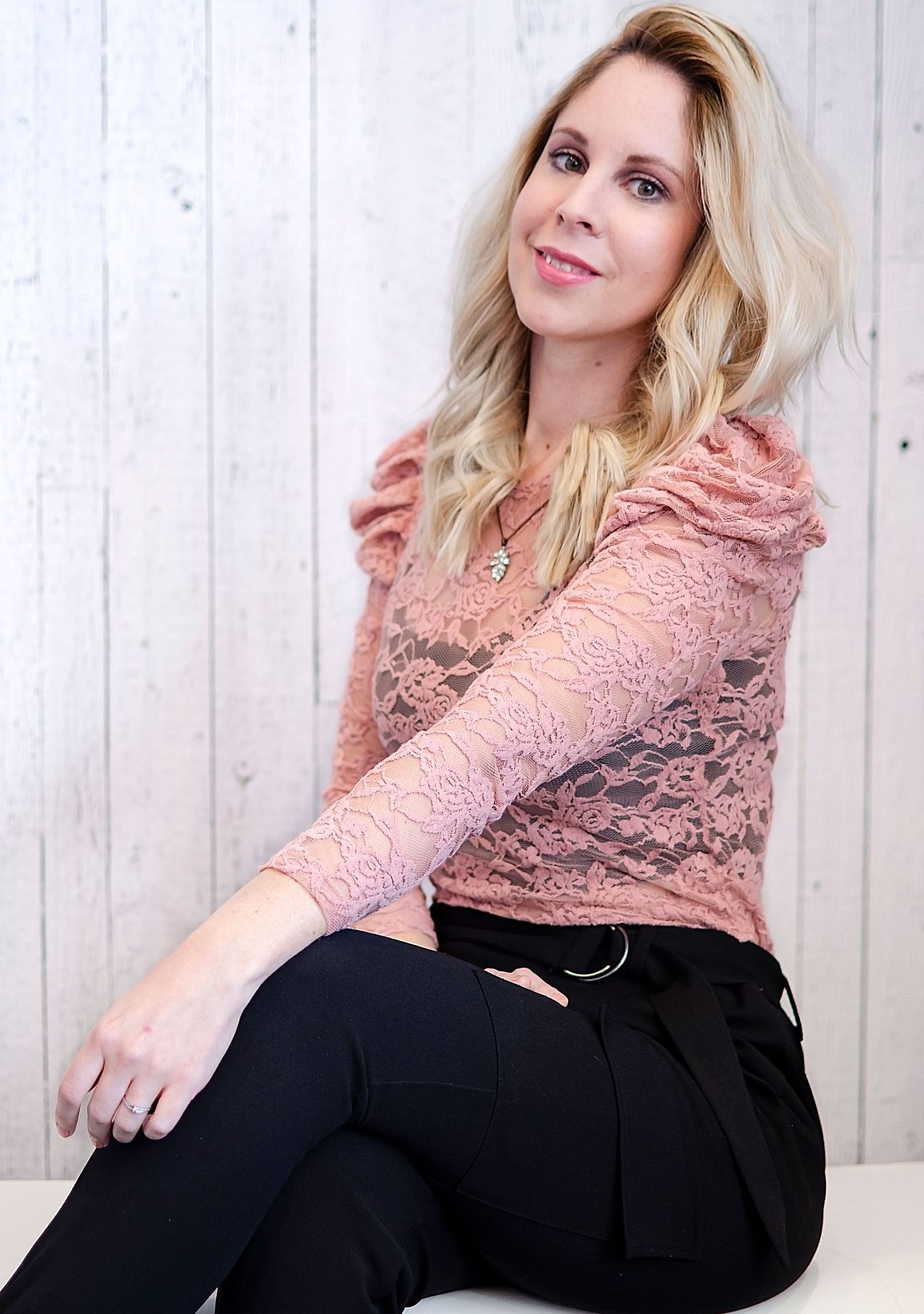 Blonde woman in pink lace top and black pants sits relaxed against white wood backdrop.