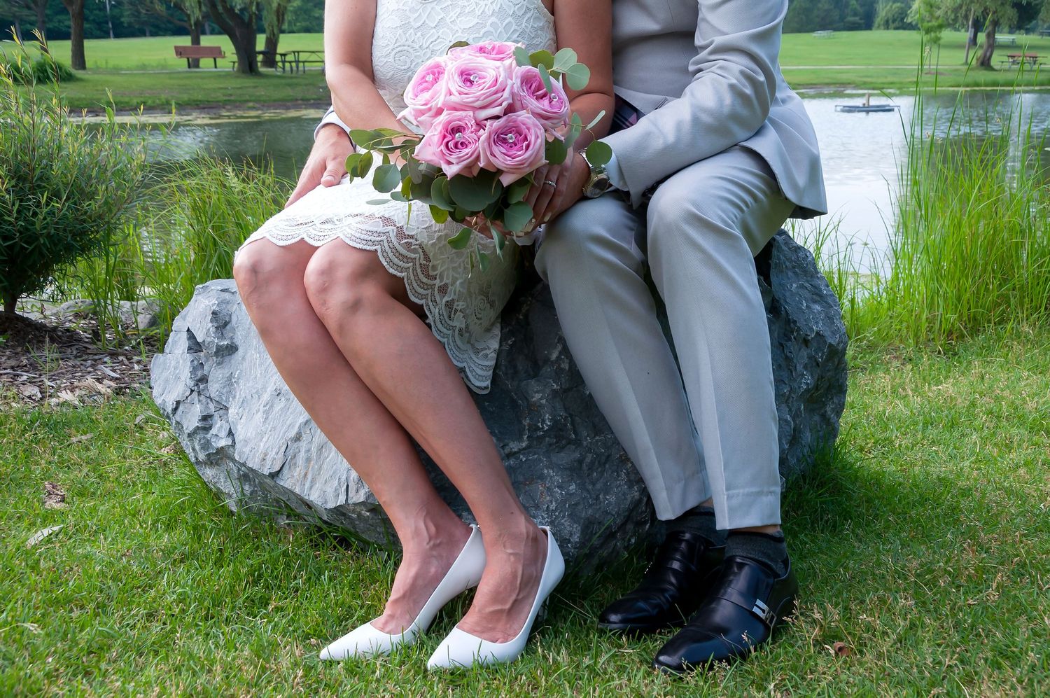 Bride and groom sitting on a boulder in a garden, bride holding a bouquet of pink roses.
