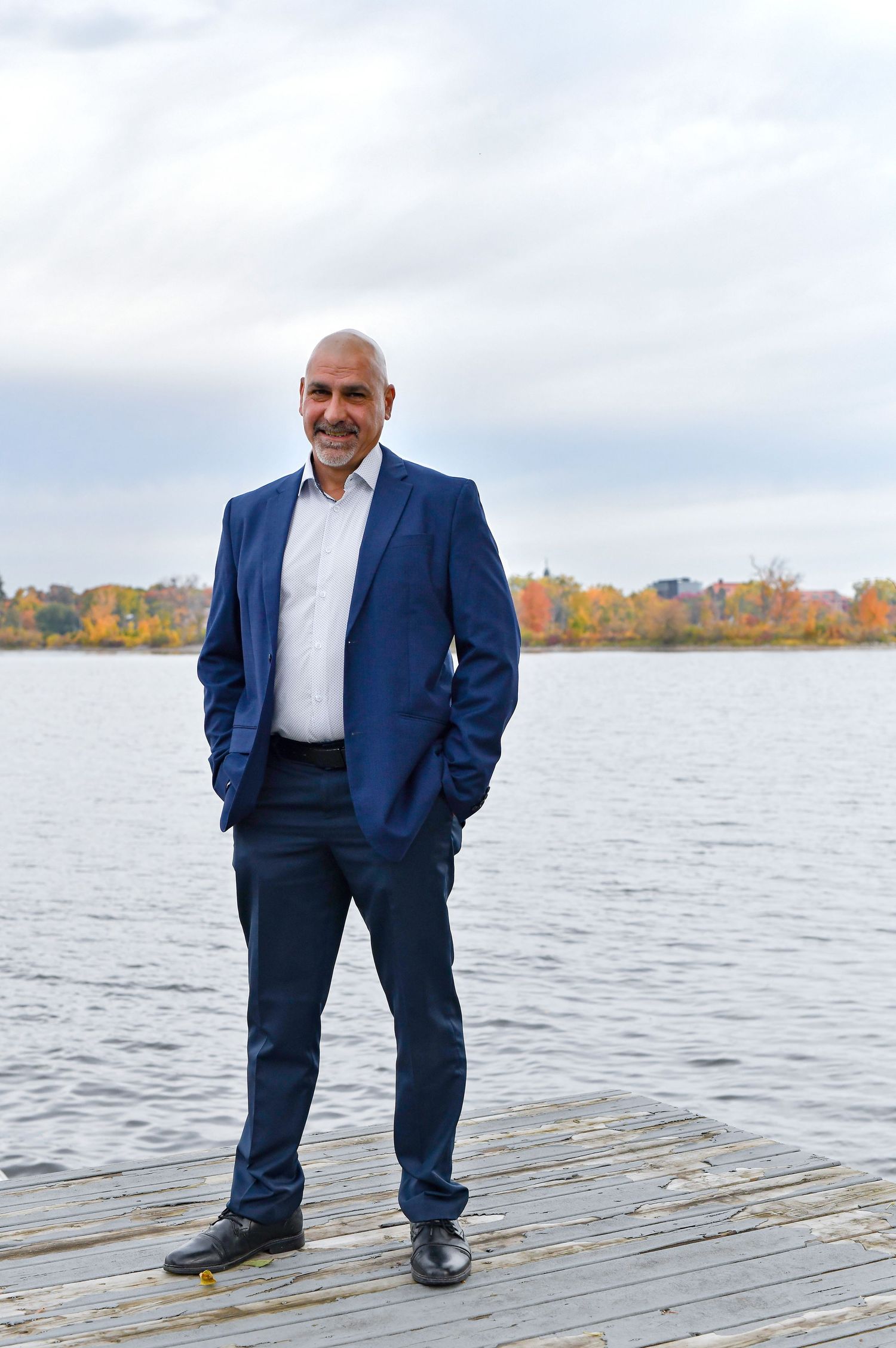 Confident man in blue suit stands on wooden dock by lake with autumn foliage backdrop.