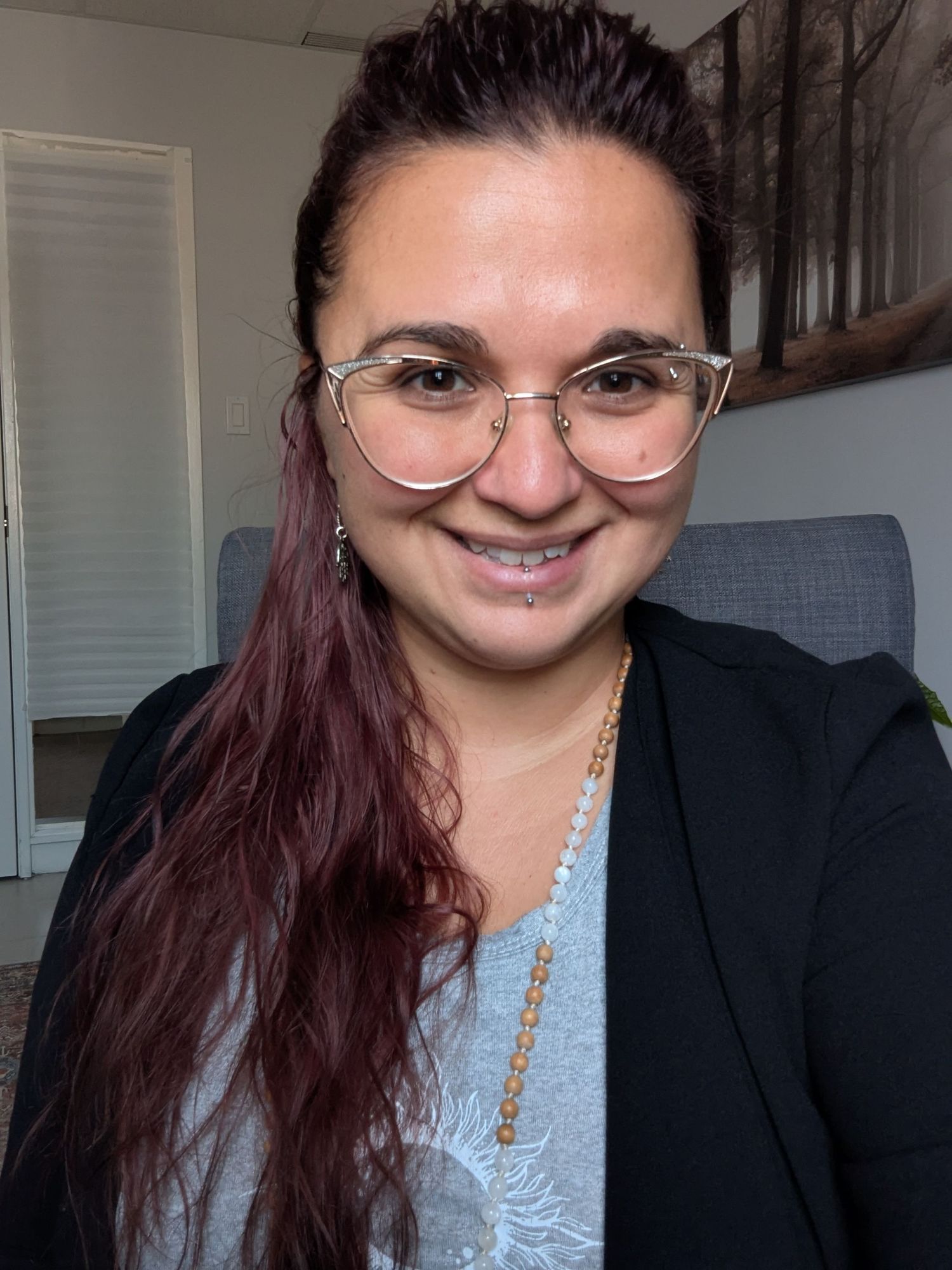 Woman with red hair and clear glasses smiles wearing black blazer and gold chain necklace indoors.