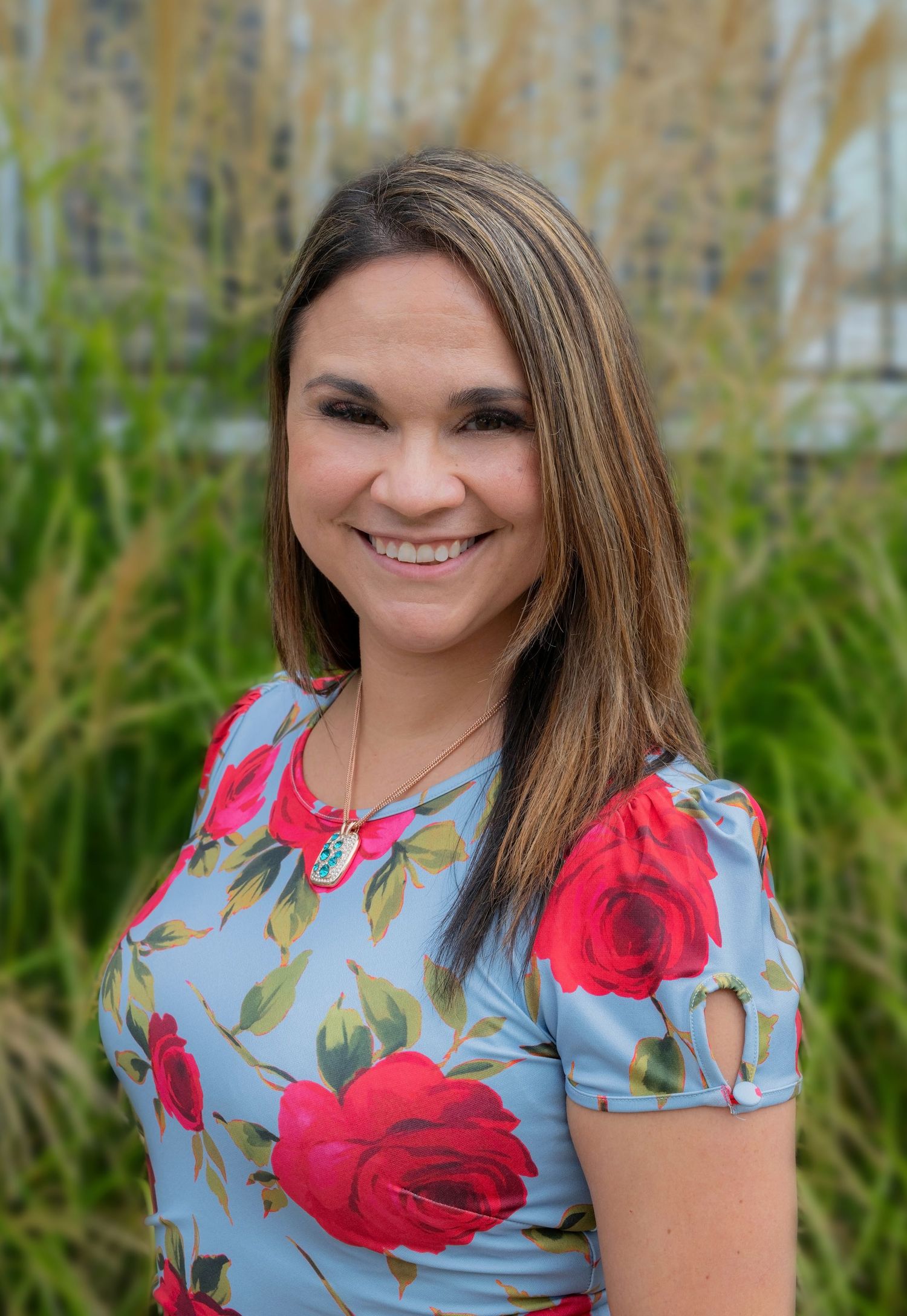 Smiling woman in floral blue dress with red roses poses outdoors near tall green grasses.