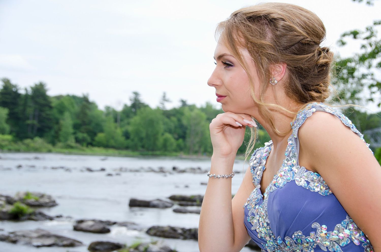 Young woman in blue floral dress gazes thoughtfully by rocky riverside with trees behind.